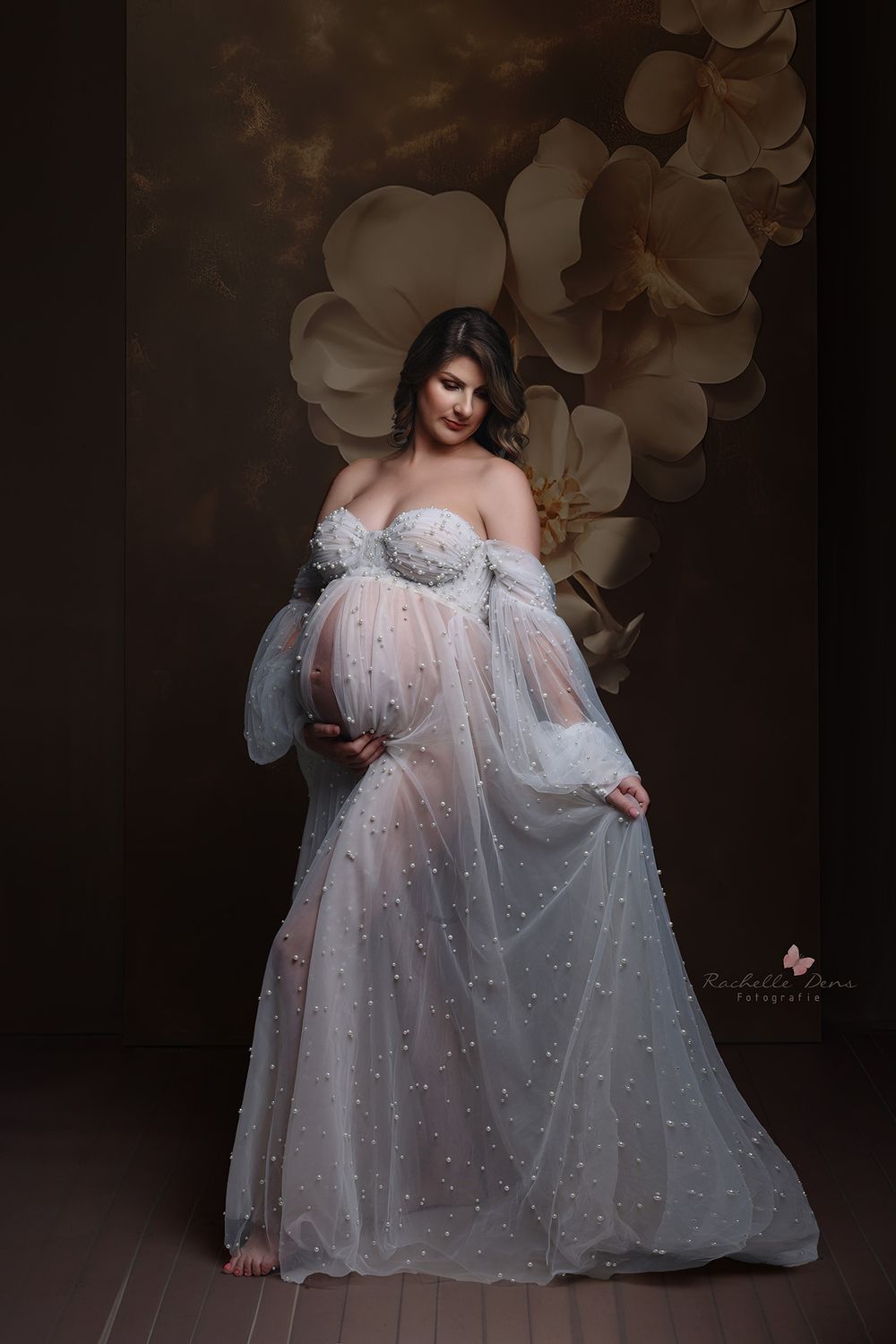 An elegant maternity portrait featuring a white off-shoulder gown with pearls against a dark backdrop with floral accents.
