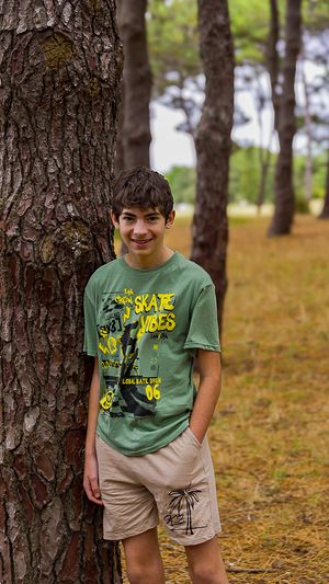 Person wearing a green t-shirt and beige shorts poses playfully among pine trees in a forest setting.