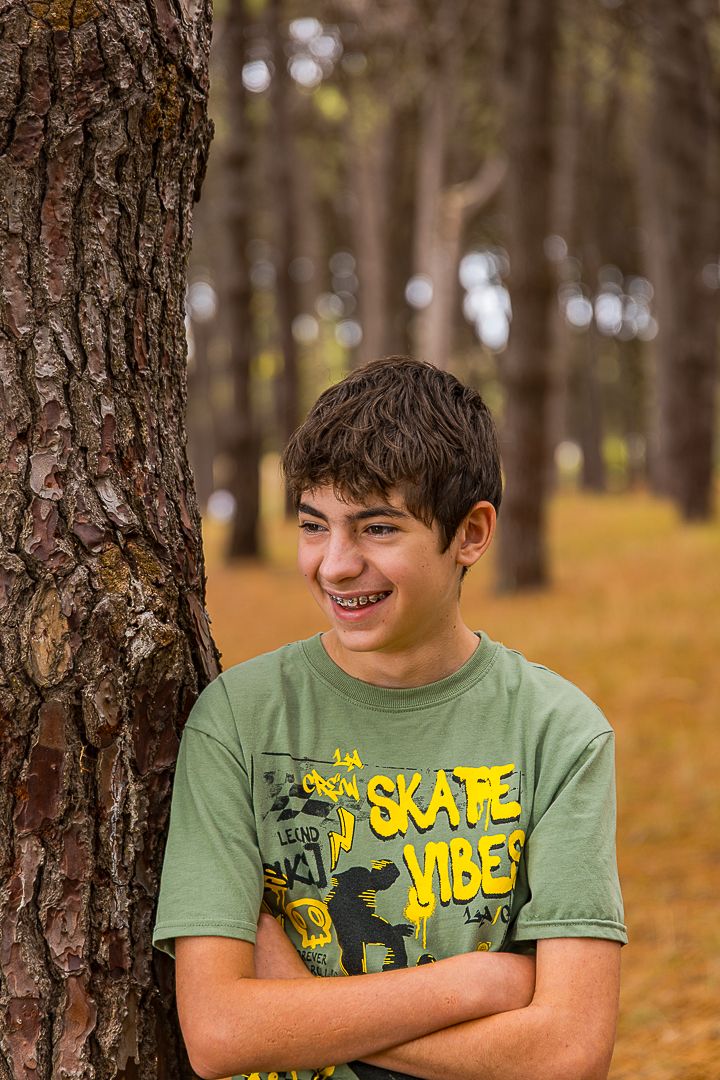 Person in a green t-shirt with yellow text sits outdoors among pine trees on an autumn day with fallen leaves.