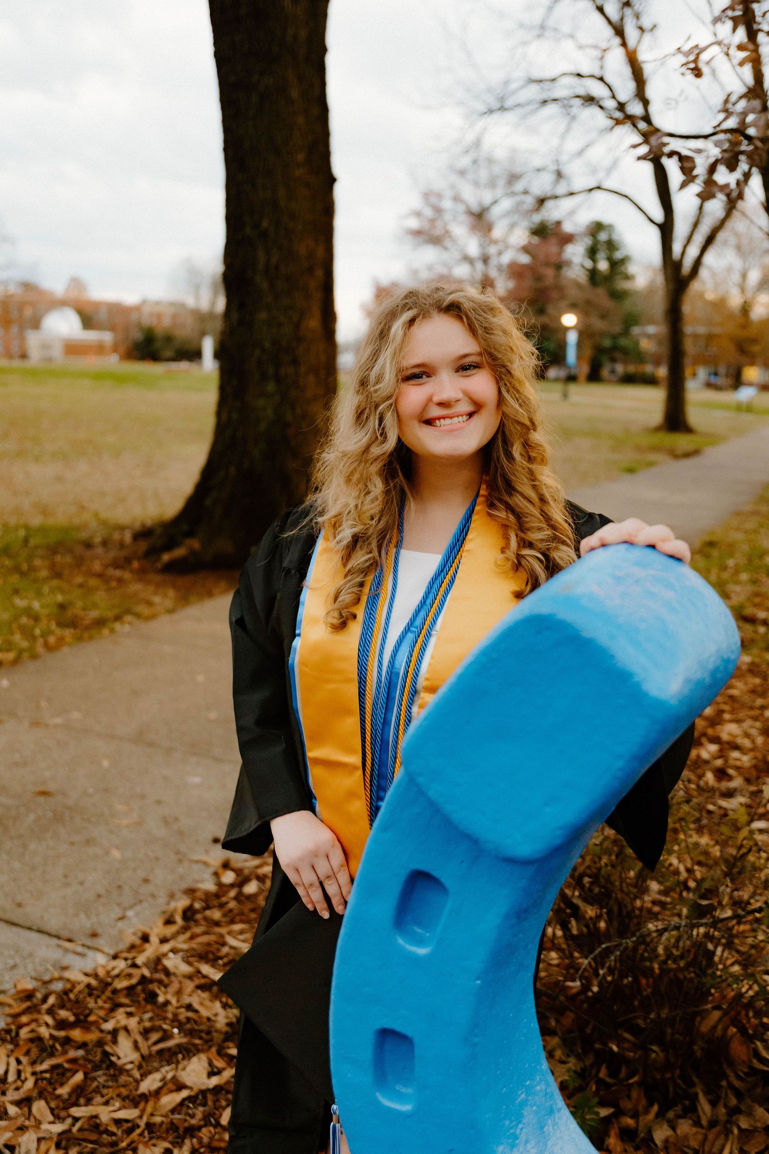 Graduate in academic regalia poses with a large blue decorative sculpture outdoors.