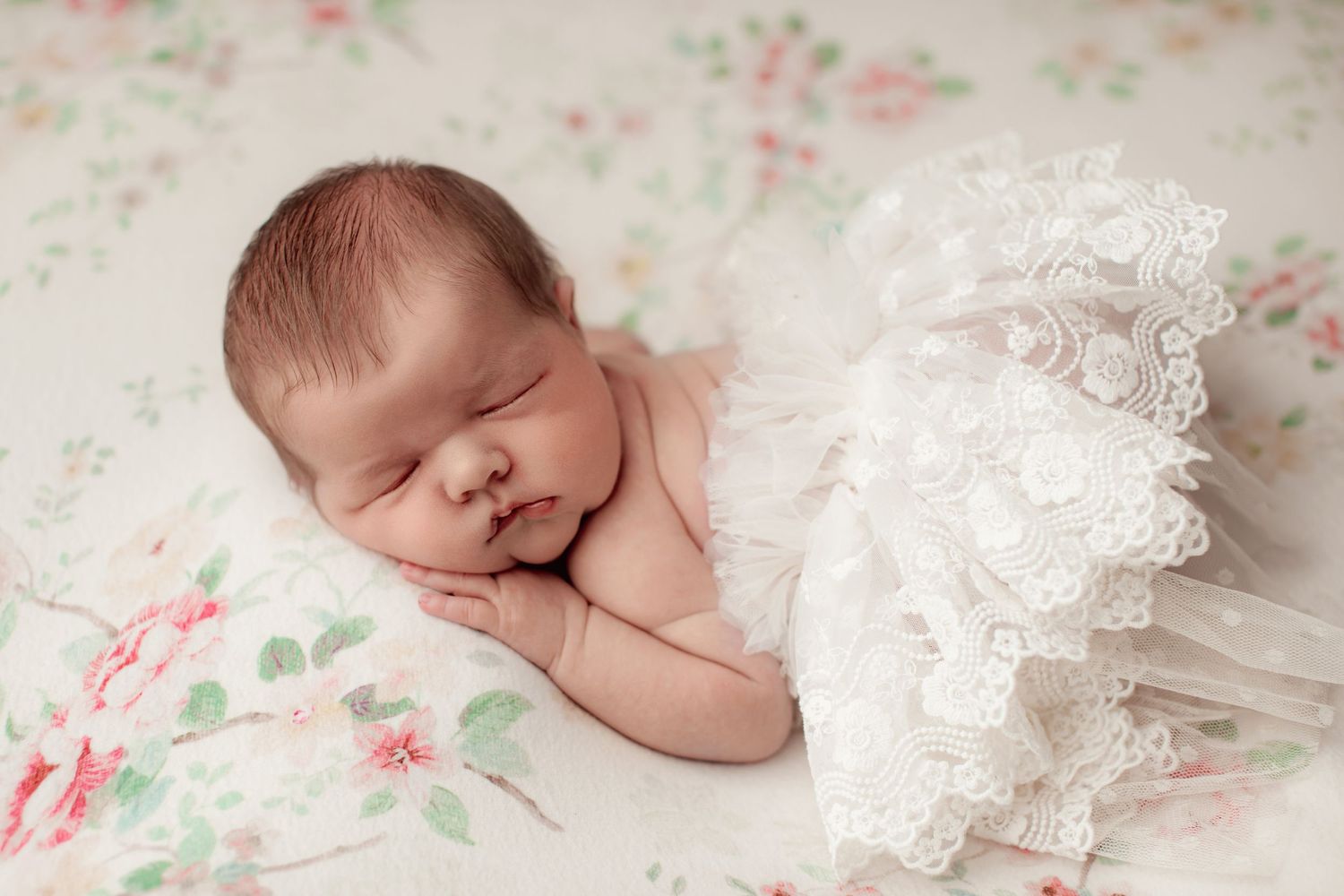 Sleeping baby lying on floral bedding, draped in delicate white lace — photographed by a professional newborn photographer Palmerston North