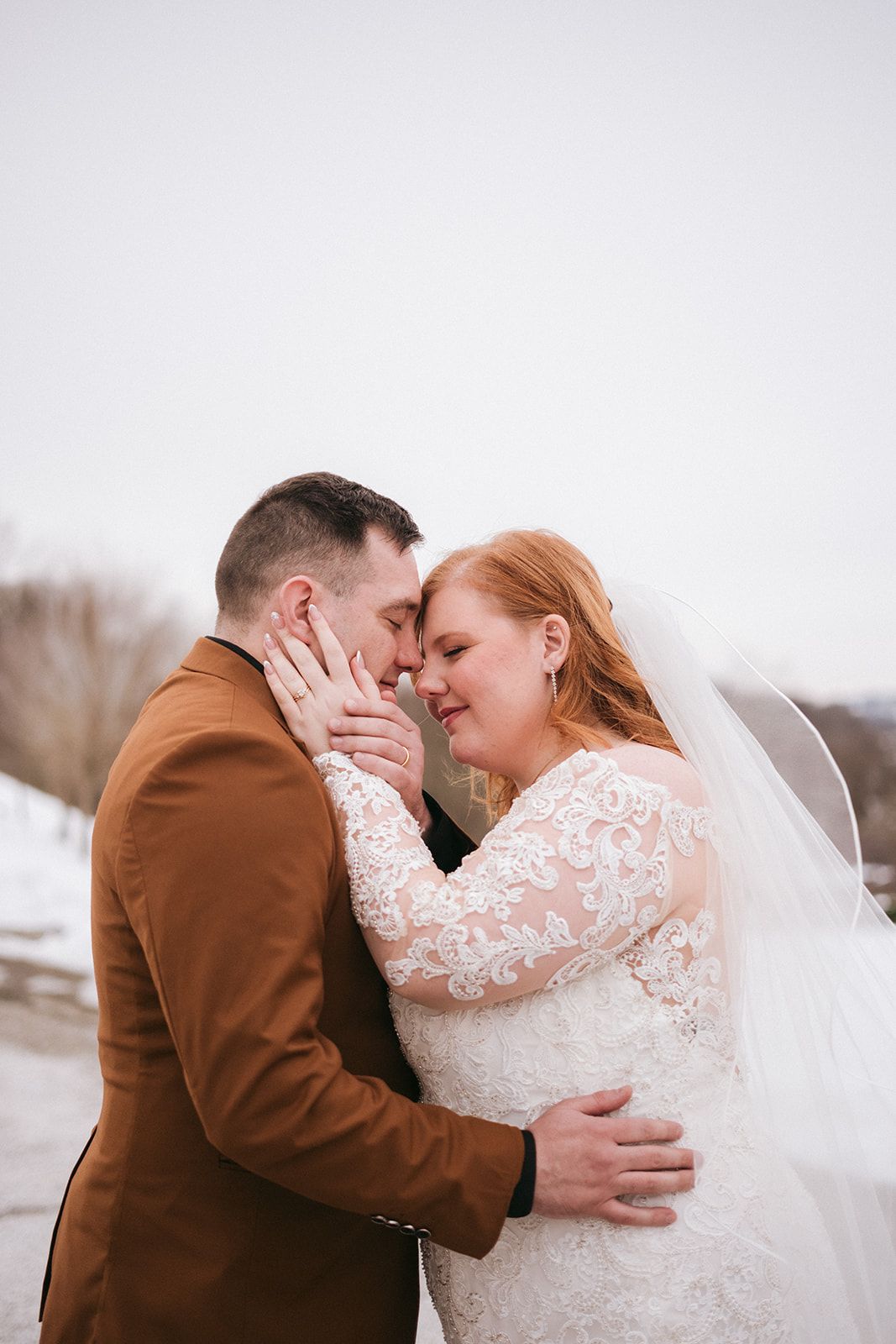 A tender winter wedding moment with the couple embracing outdoors, bride in lace and groom in brown suit.