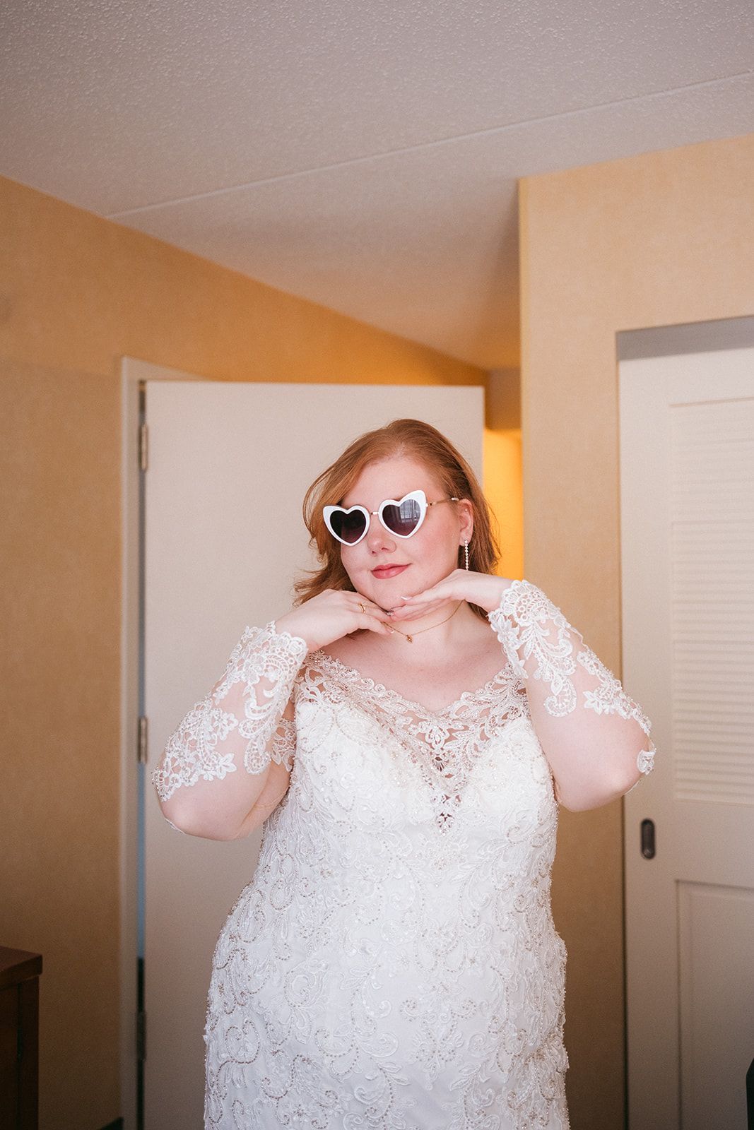 A person wearing a white lace wedding dress and heart-shaped sunglasses poses playfully in a room.
