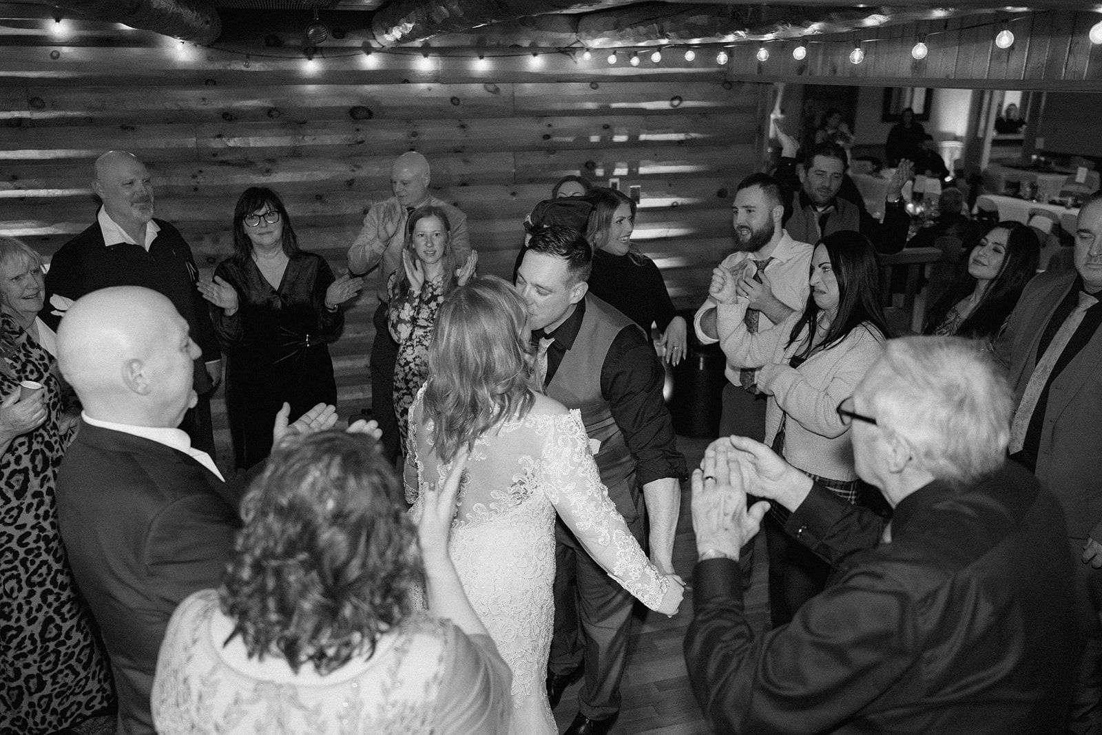 A group celebrates together on a dance floor at a wedding reception, captured in black and white photography.