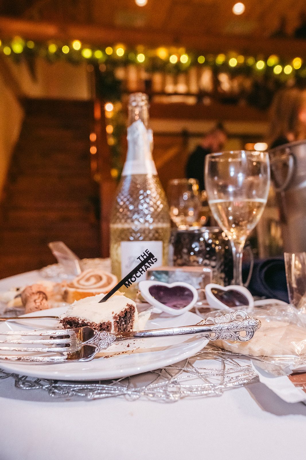 A romantic table setting with wine glasses, heart-shaped sunglasses, and a decorative bottle with ambient string lights.