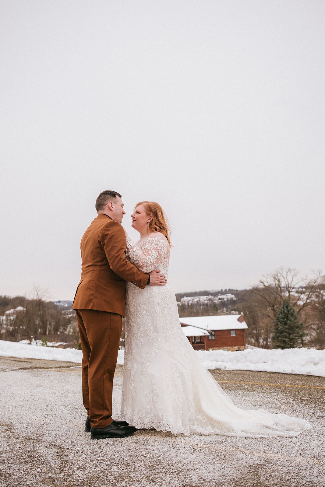 A couple embraces outdoors in winter with snow on the ground, bride wearing white lace and groom in brown suit.