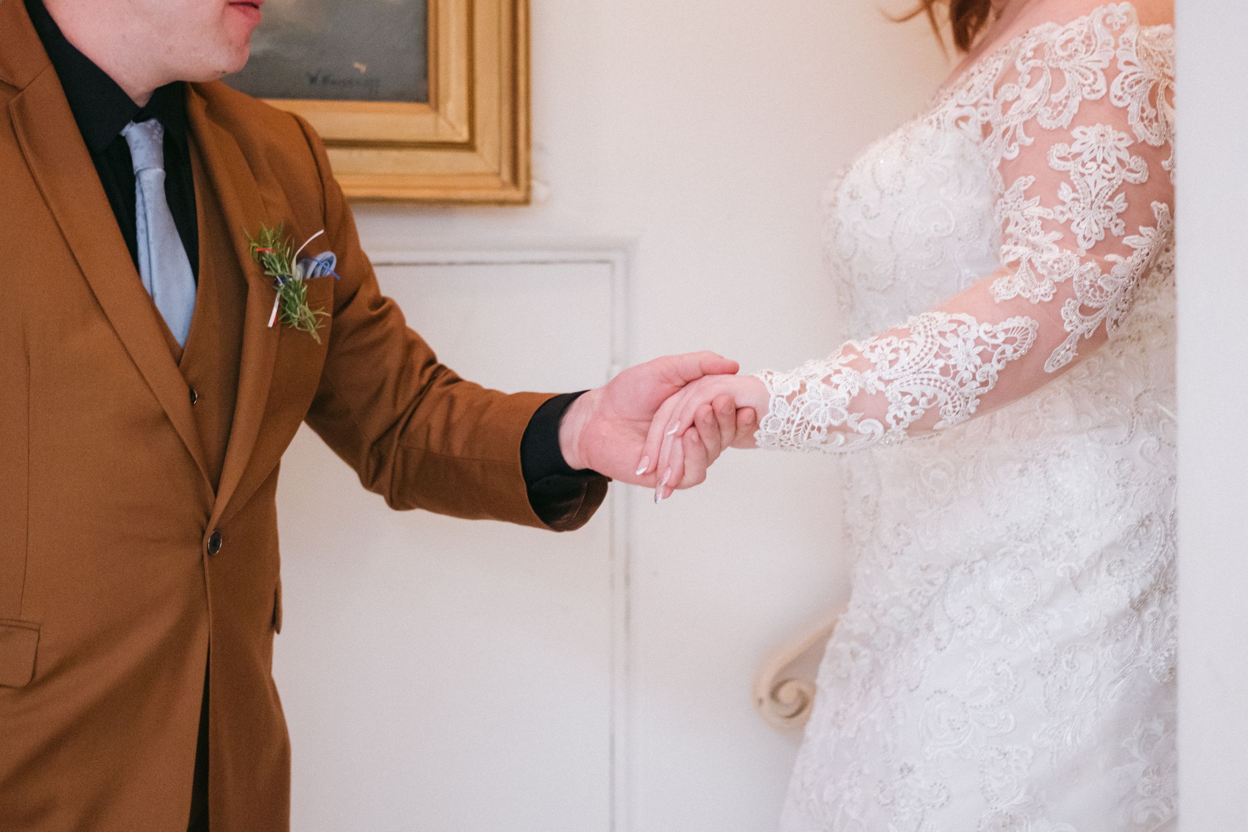 A close-up of wedding attire showing a brown suit jacket and white lace dress sleeve as hands join together.