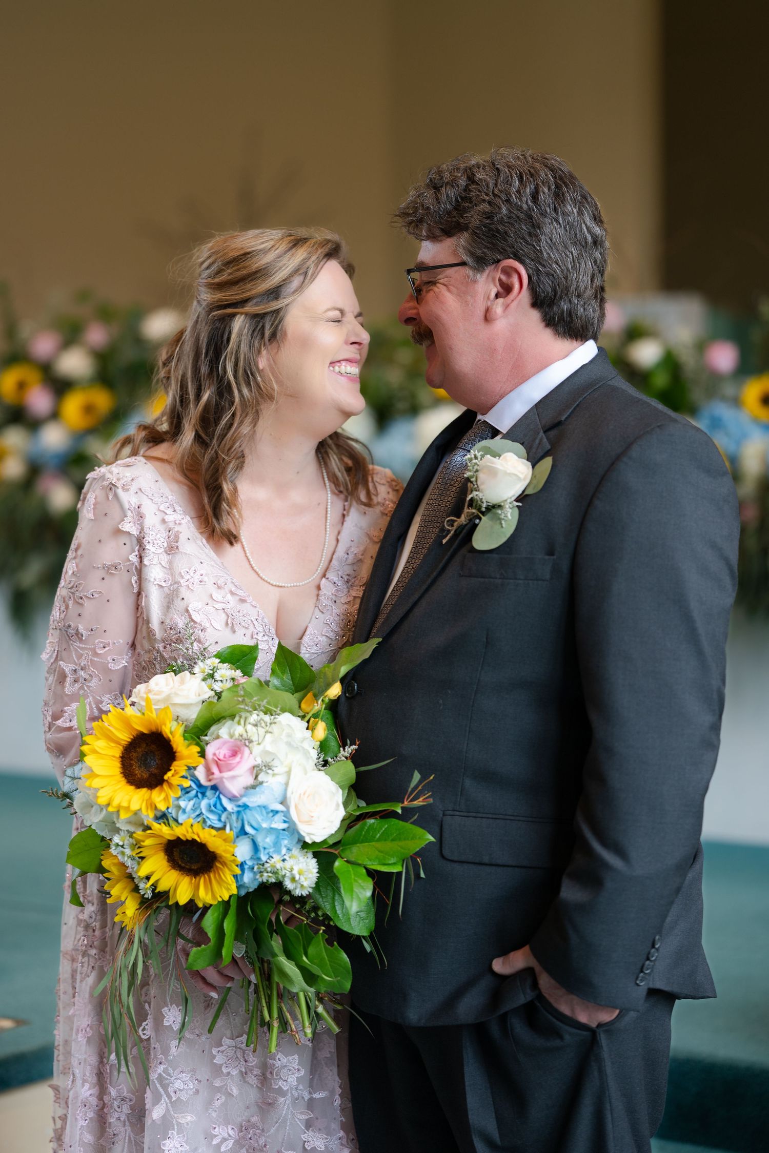 Wedding couple share a joyful moment as bride holds vibrant sunflower bouquet in her lace gown beside groom in dark suit.