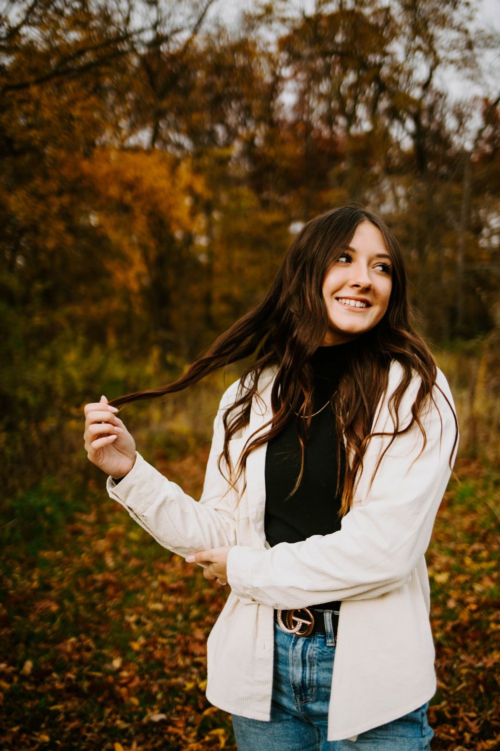 A person in a white jacket and jeans poses playfully against an autumn background with fallen leaves.