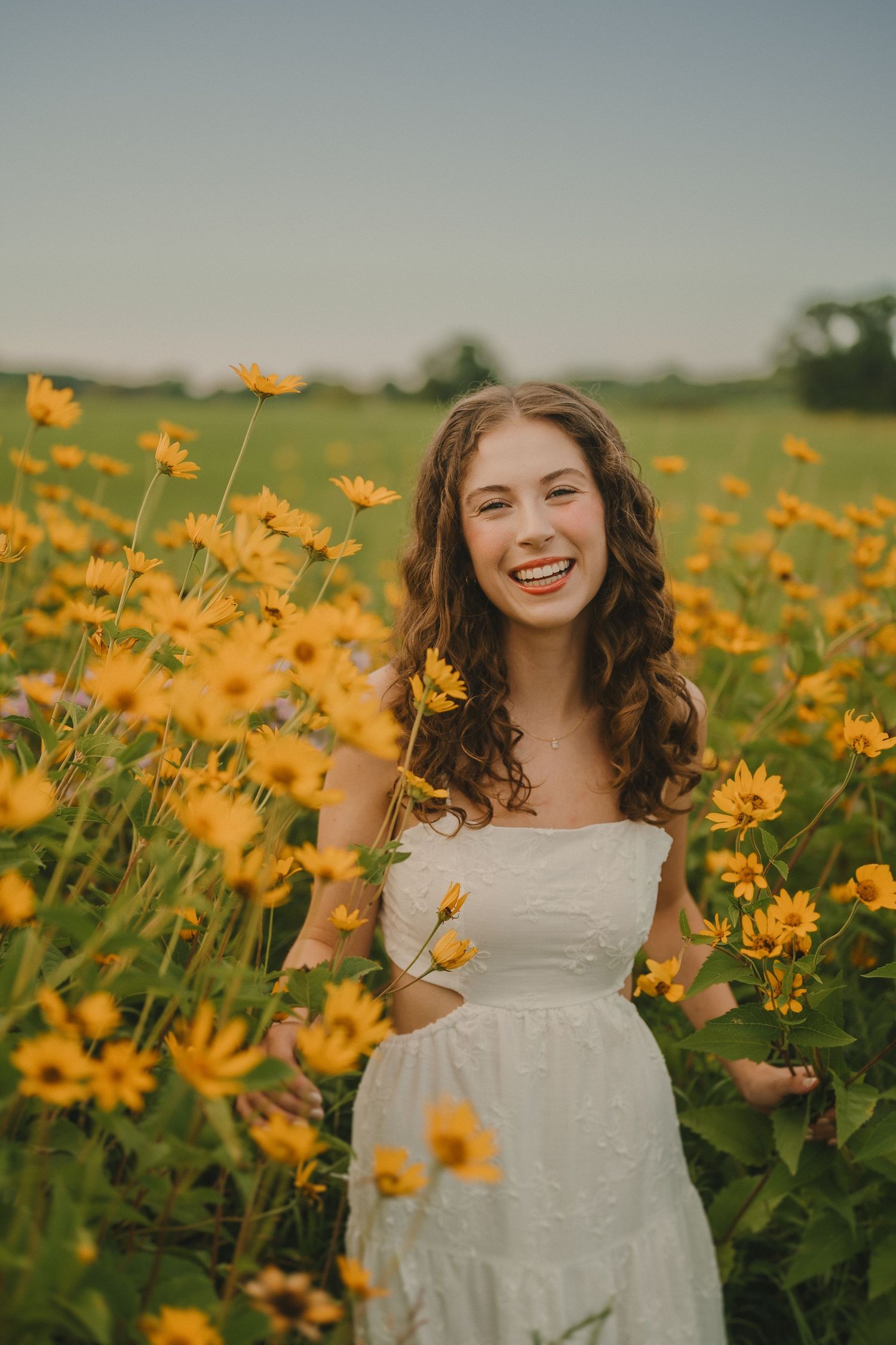 A white sundress-clad woman stands joyfully among vibrant yellow wildflowers in a sunlit meadow.