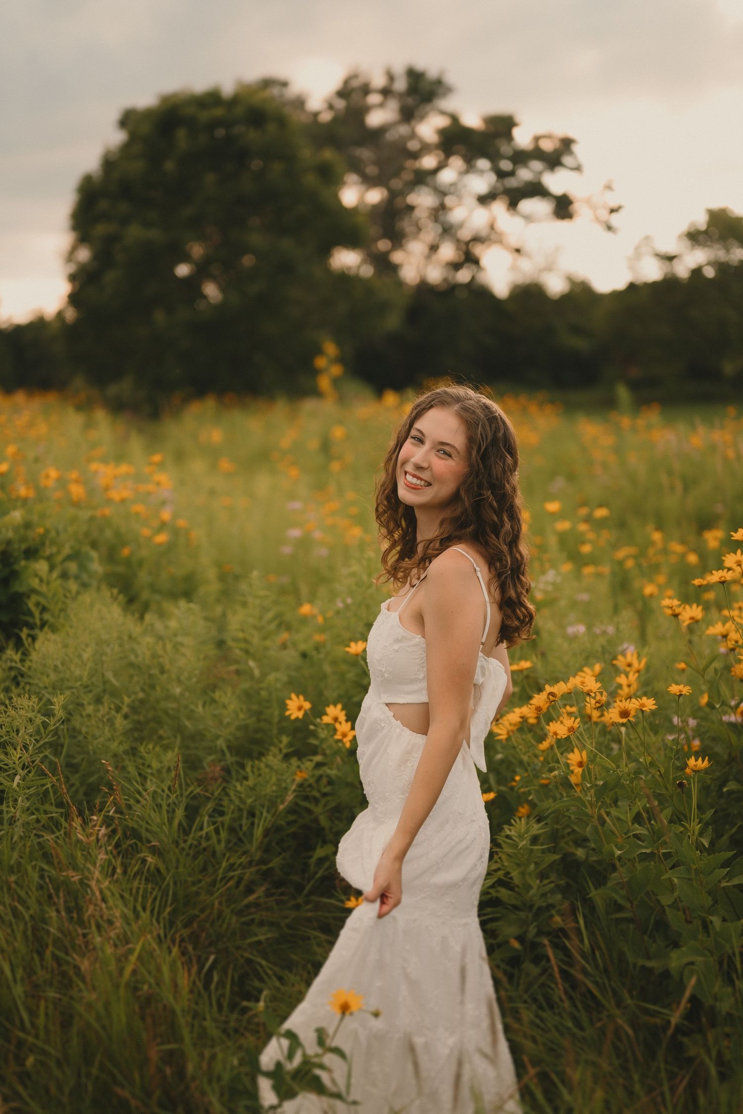 A white dress flows elegantly as someone poses in a golden meadow at sunset with wild daisies.
