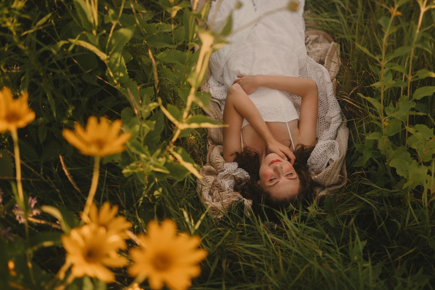 A dreamy portrait of a person lying in tall grass surrounded by bright yellow sunflowers on a summer day.