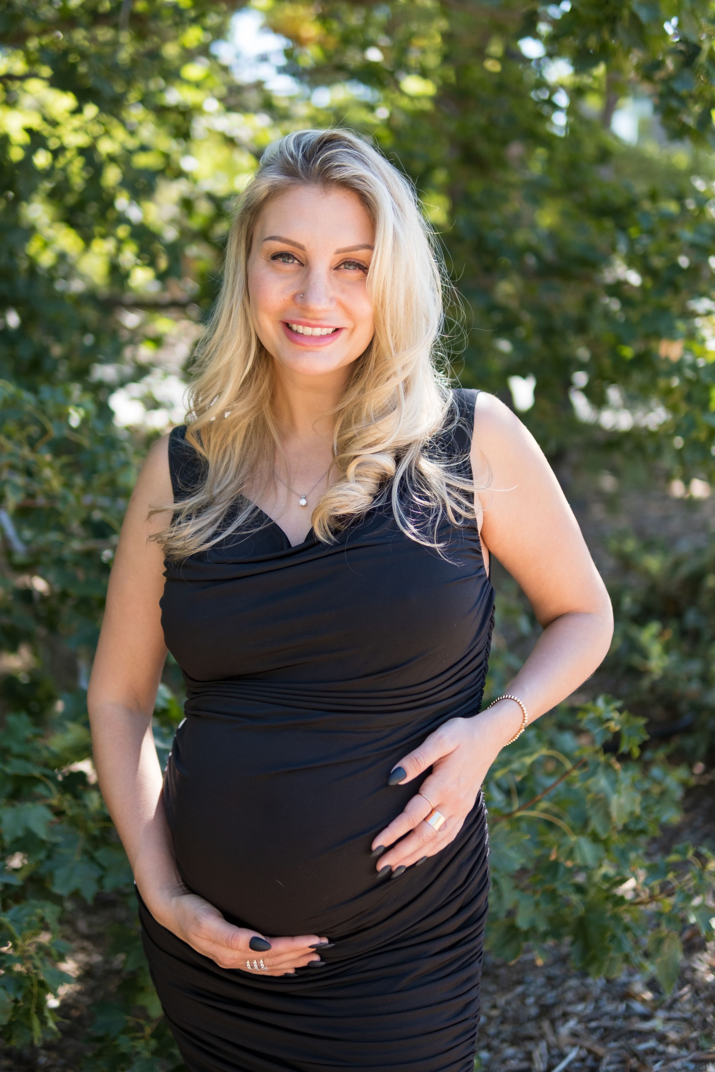 Person in black sleeveless maternity dress stands outdoors with sunny foliage background during pregnancy photo session.