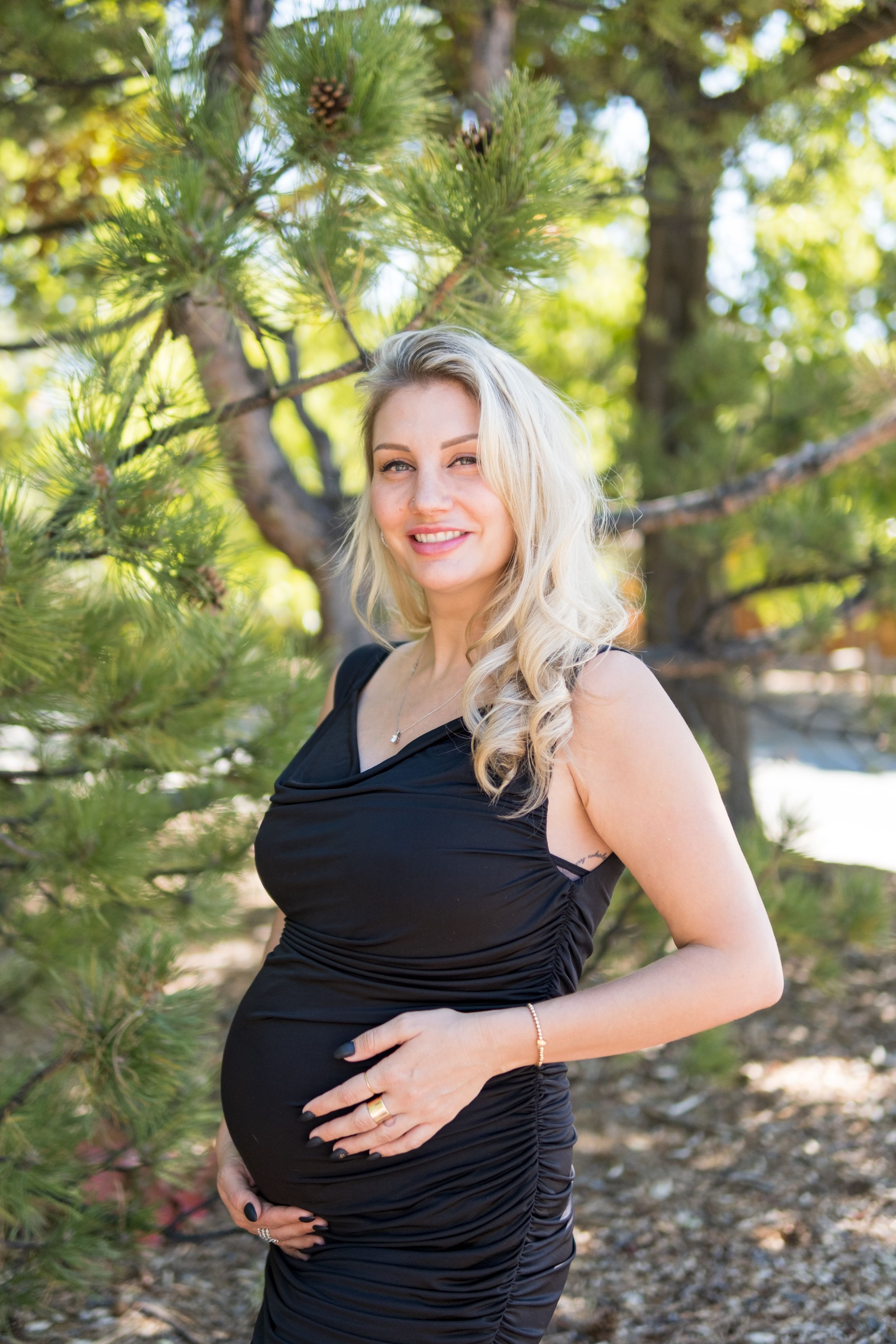 Expectant woman in fitted black dress poses outdoors among pine trees in natural sunlit setting.