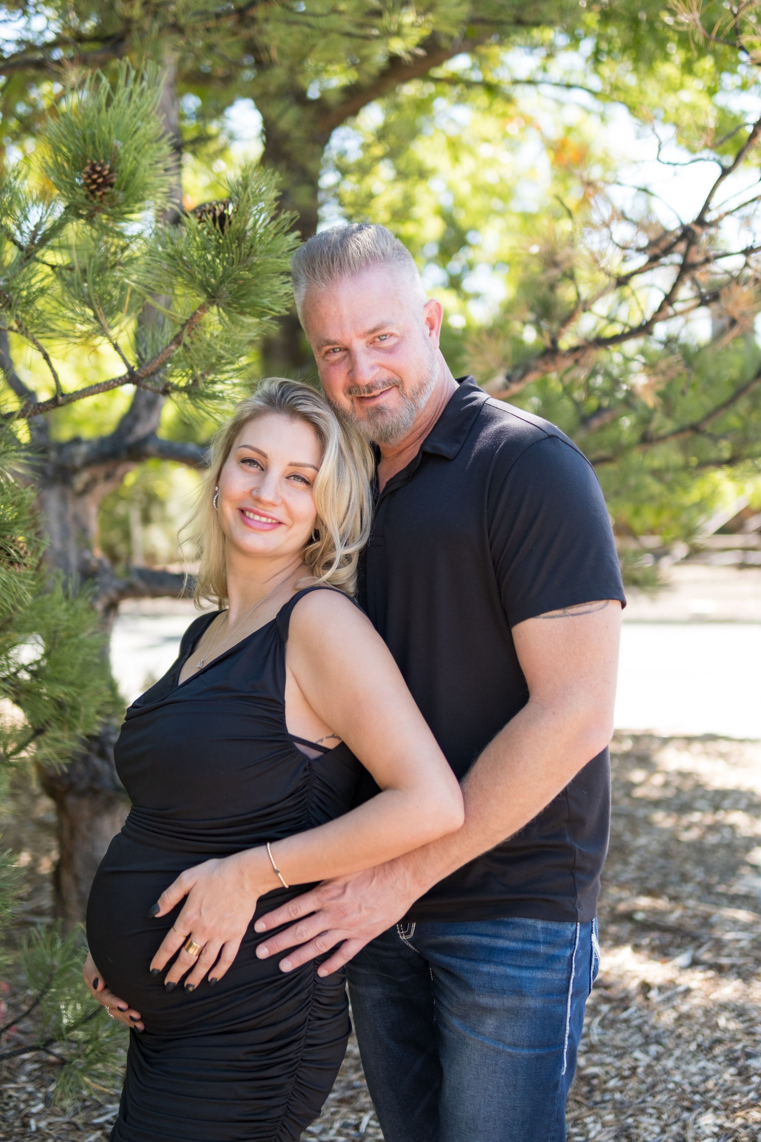 A couple poses together in a wooded outdoor setting with sunlight filtering through pine trees.