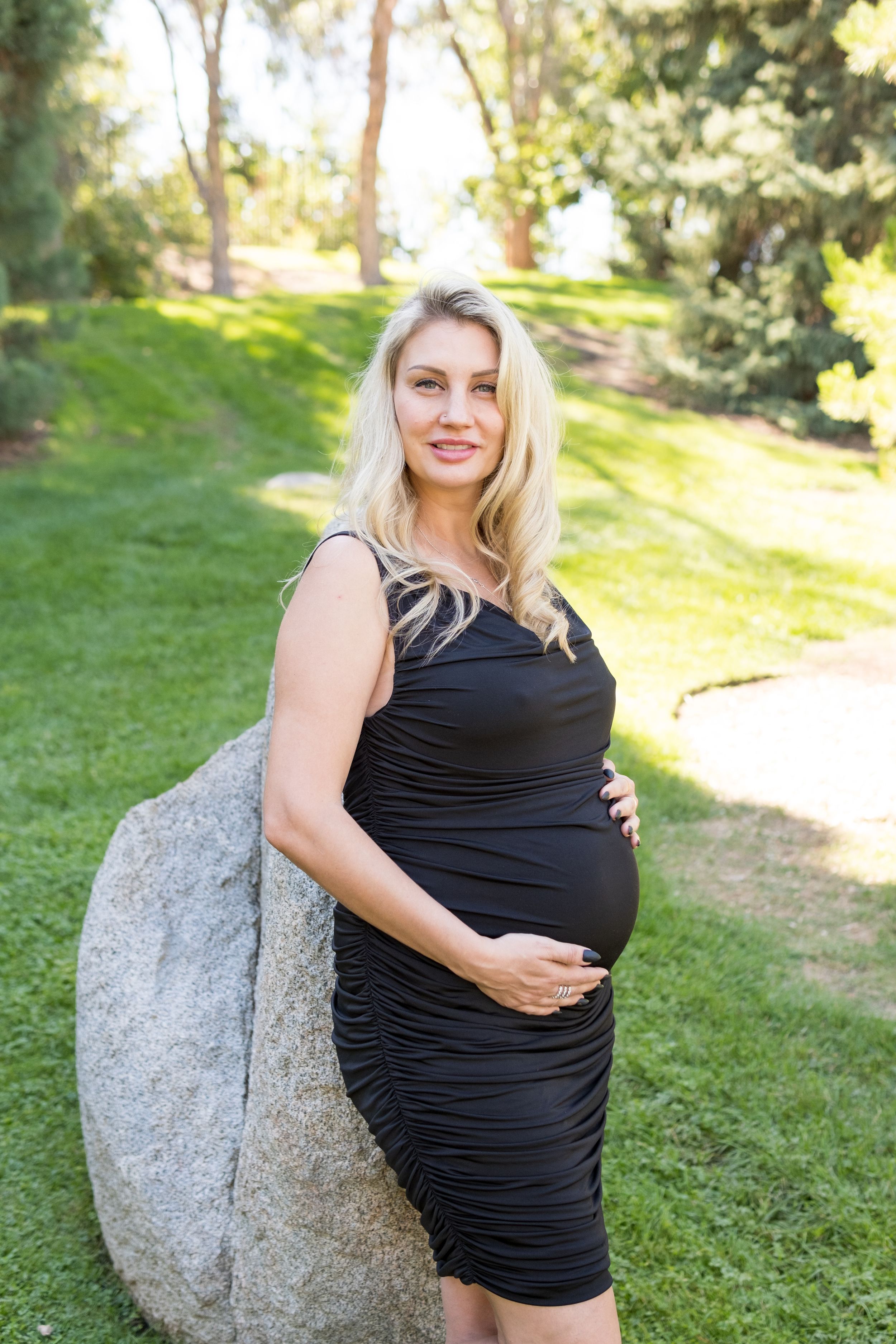 A pregnant woman in a black ruched dress poses outdoors in a sunny garden setting next to a large boulder.