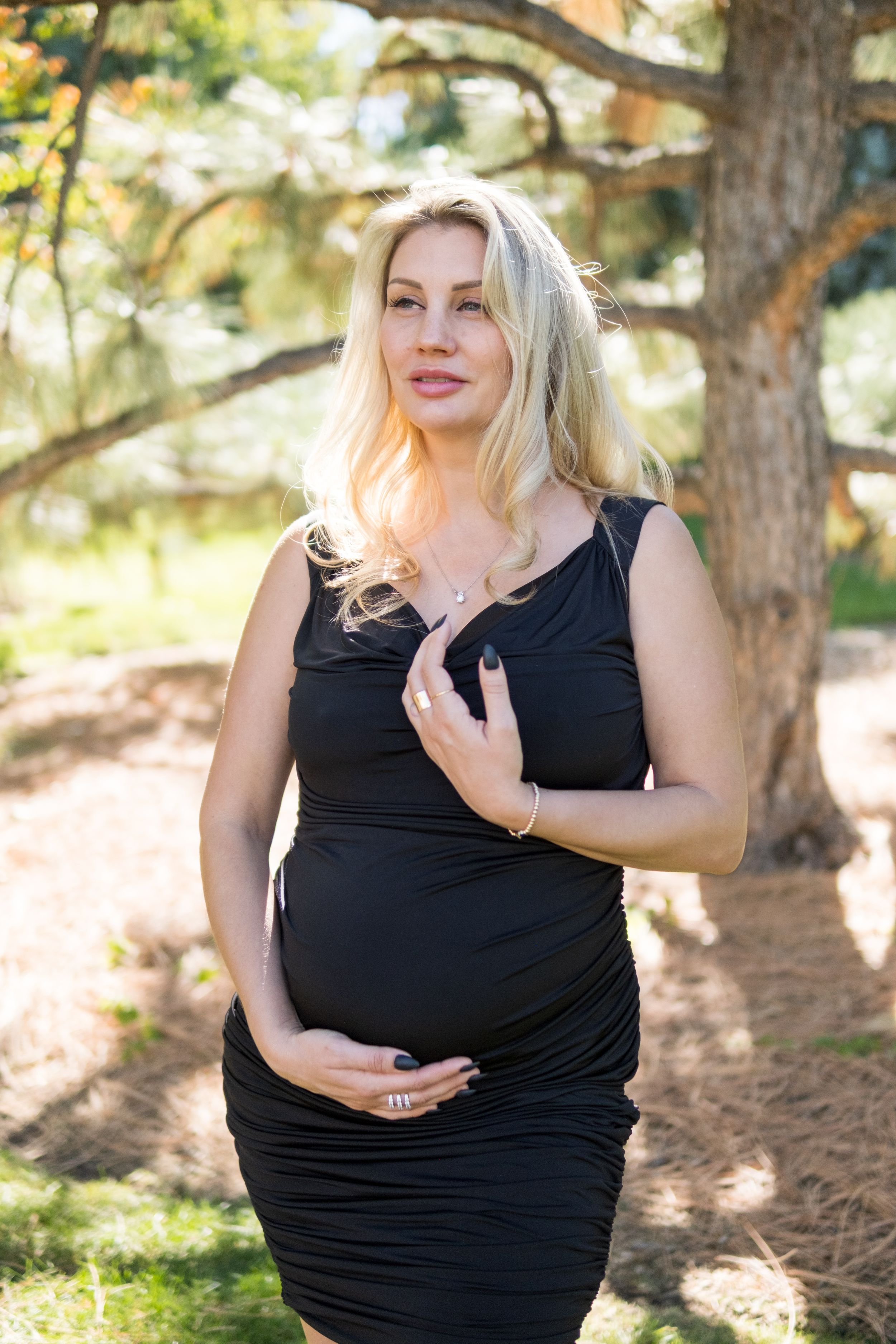 A pregnant woman in a black dress poses outdoors against a natural wooded backdrop with soft lighting.
