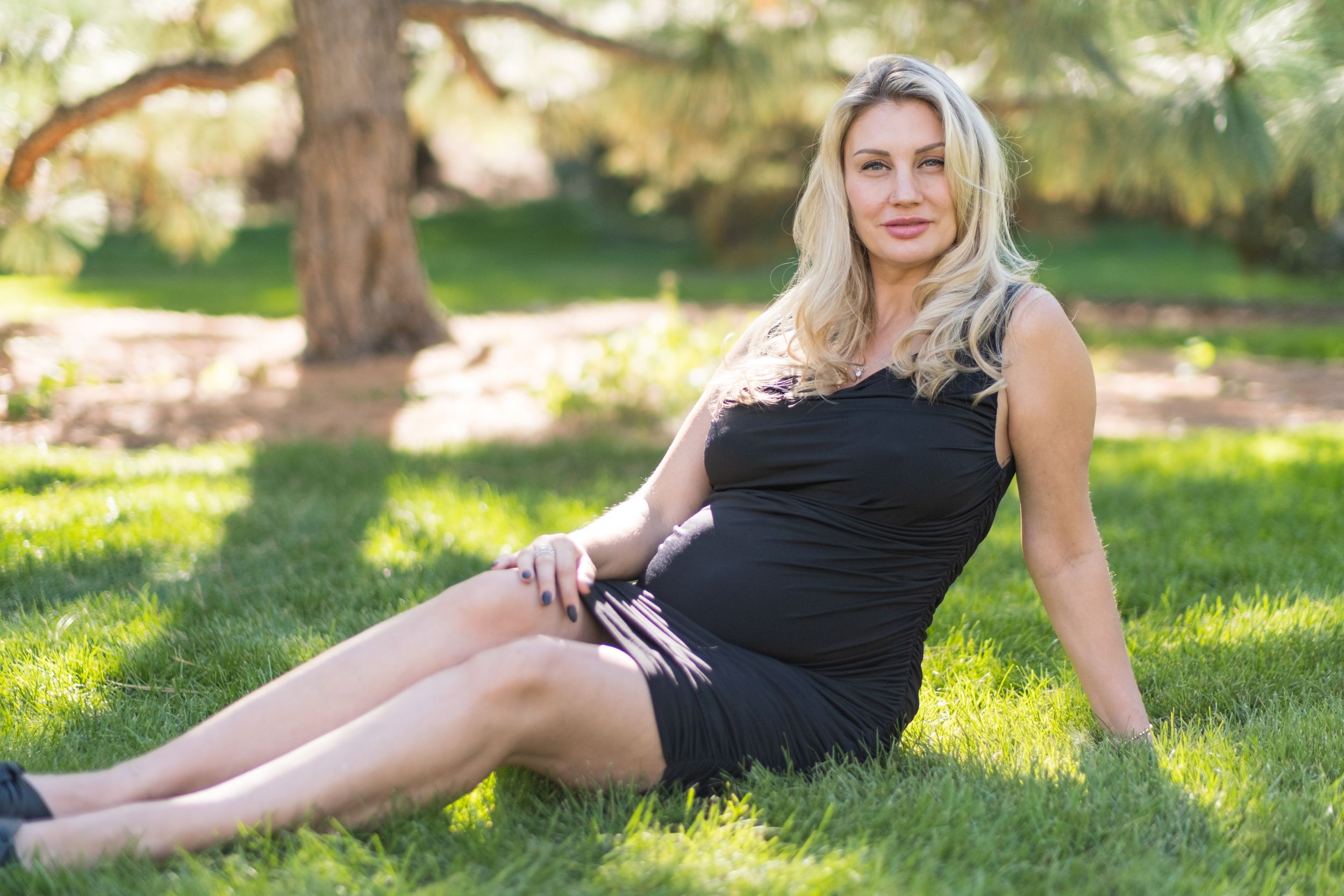 A woman in a black dress lounging on grass in a sun-dappled park setting during summer.
