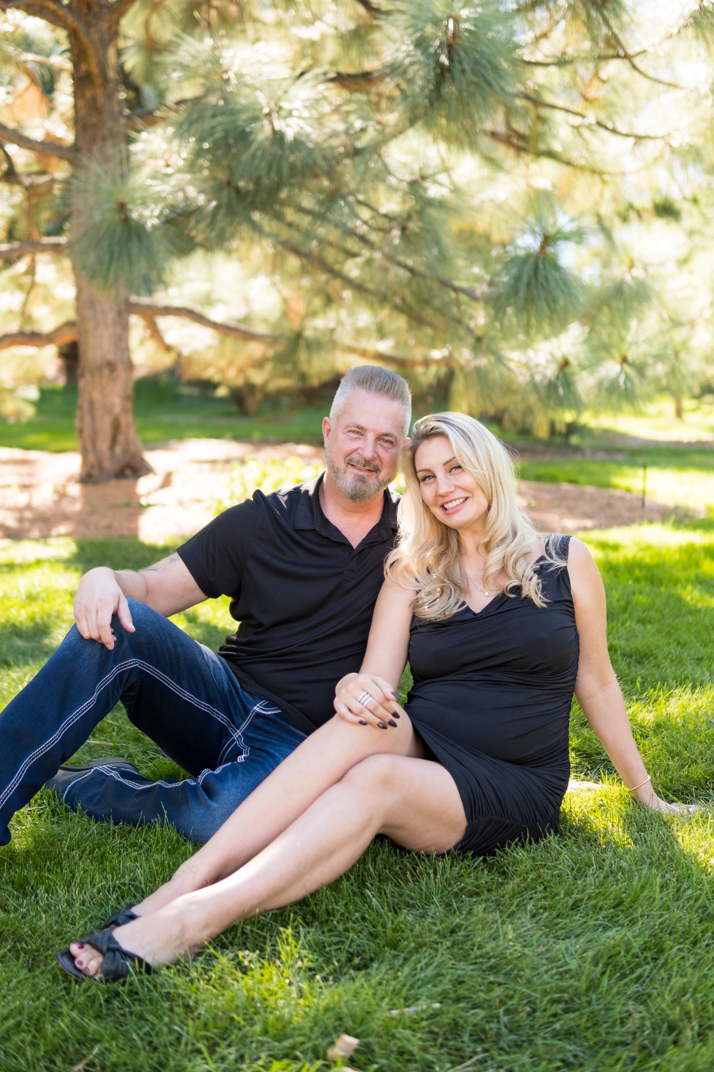 A couple sits together on green grass beneath pine trees on a sunny afternoon in a park setting.