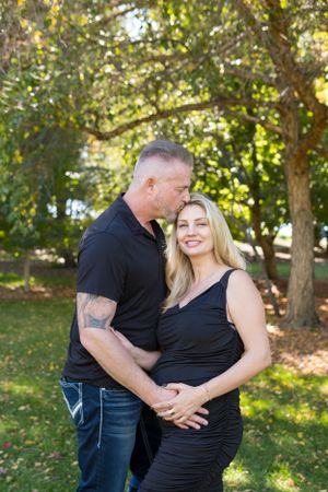 A couple wearing black clothing embraces in a sunny garden setting with trees in the background.