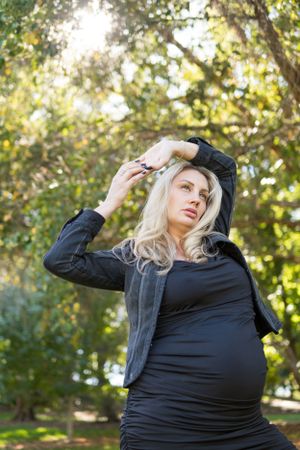 Pregnant woman in all black attire poses outdoors with sunlit trees in the background on an autumn day.