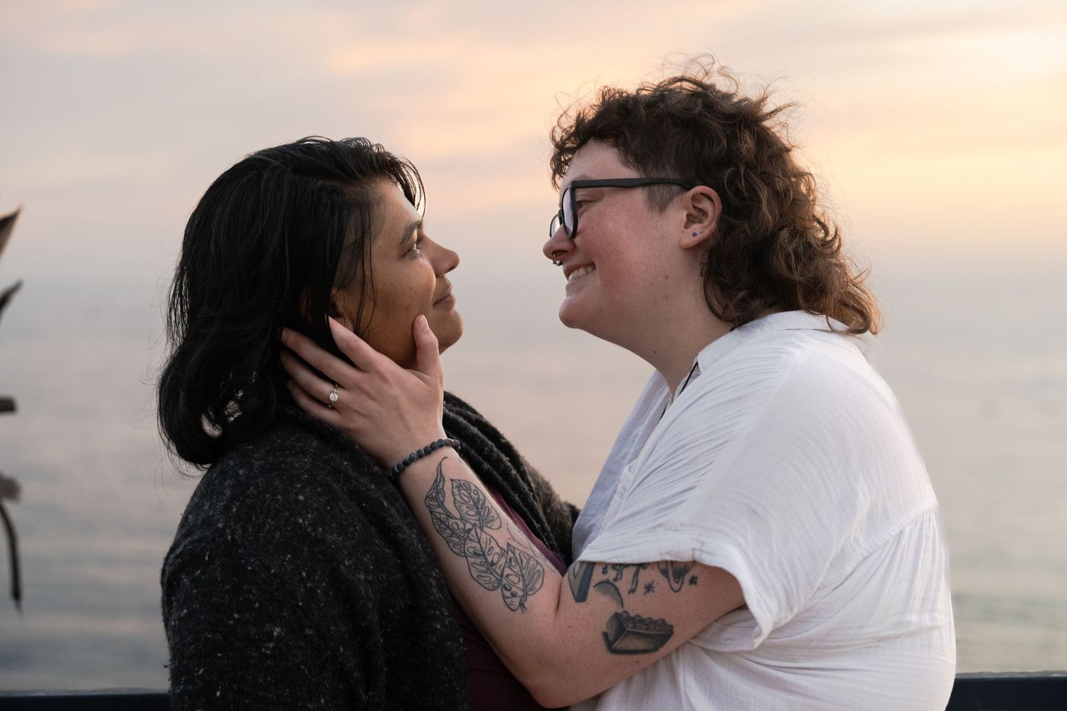 A woman loving woman couple gazes into each other's eyes with one's hand on the other after getting engaged with the ocean in the background