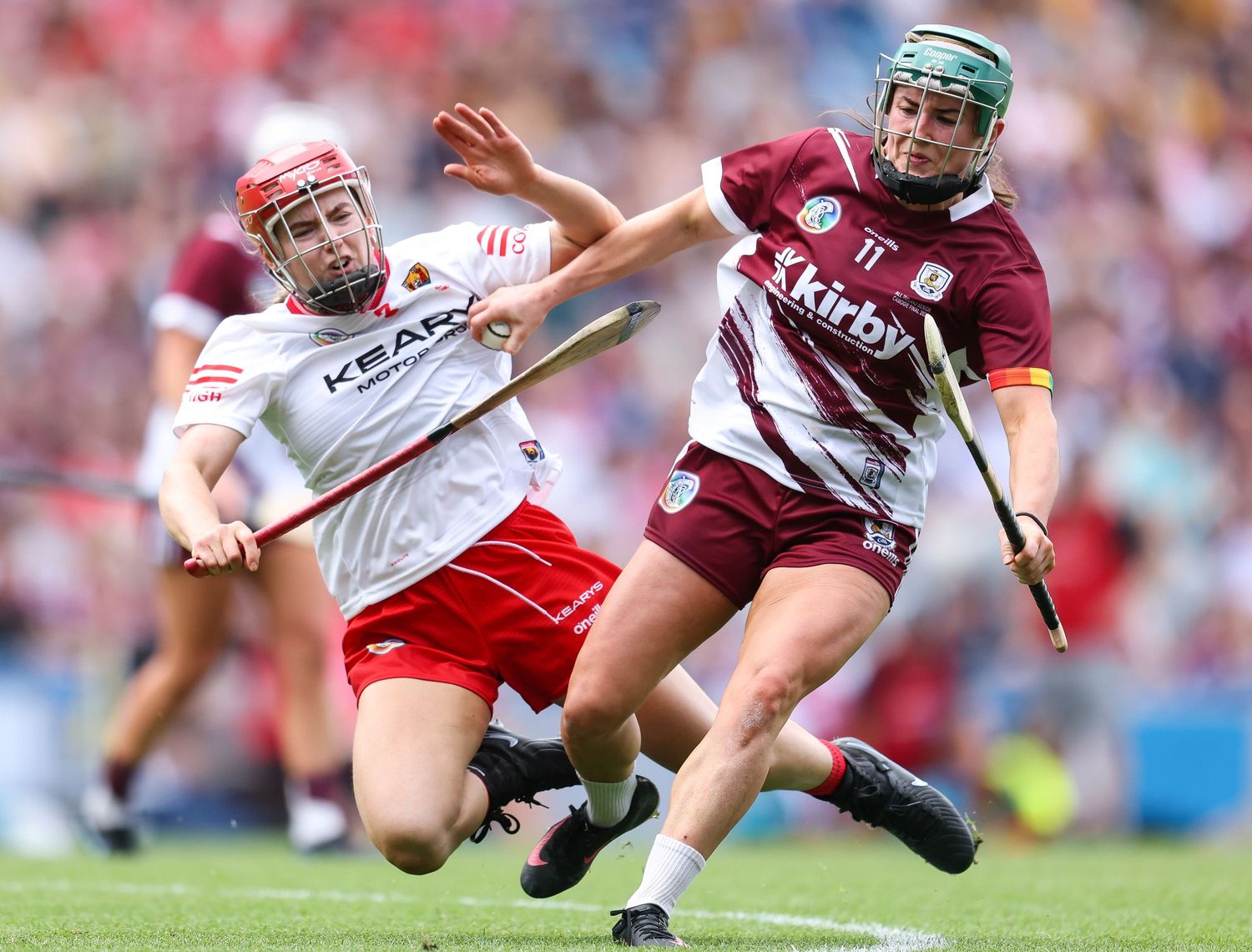 Two camogie players compete fiercely during a match, one in red and white, the other in maroon and white jerseys.