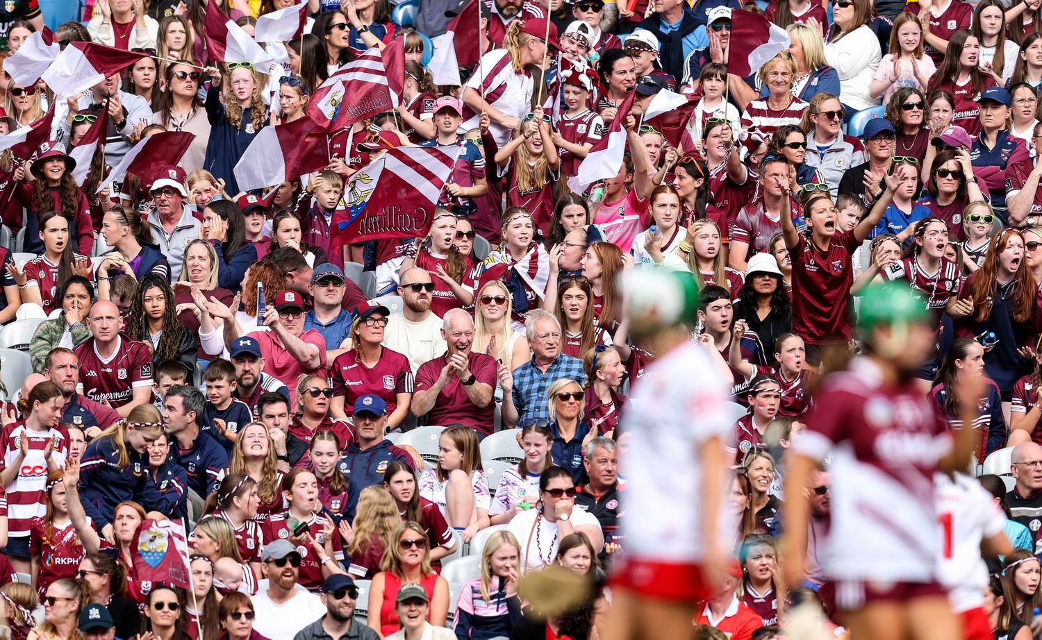 Large crowd of Galway GAA fans in maroon jerseys cheering at a Gaelic football match on a sunny day.