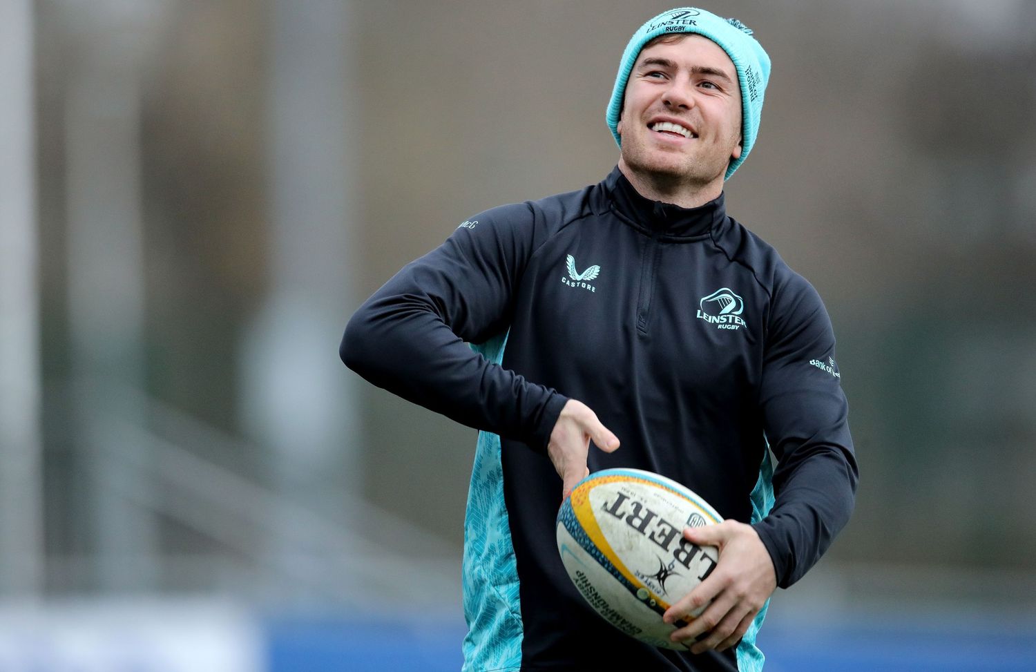Rugby player in teal beanie and black training gear holds ball during practice.