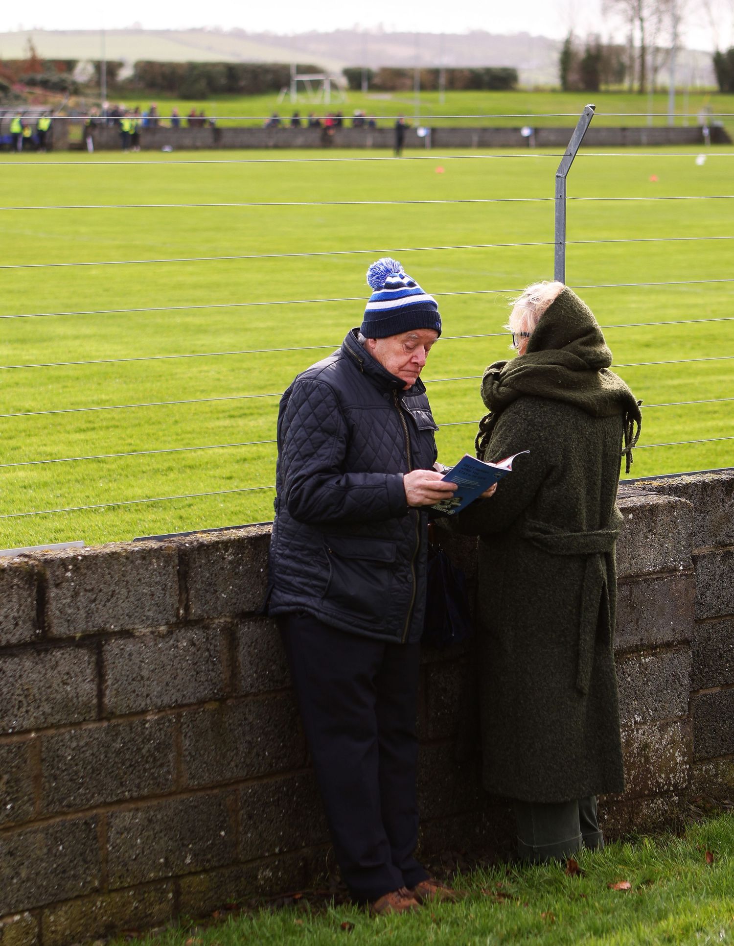 Person in blue jacket sits on stone wall while looking at mobile phone in grassy field on cloudy day.