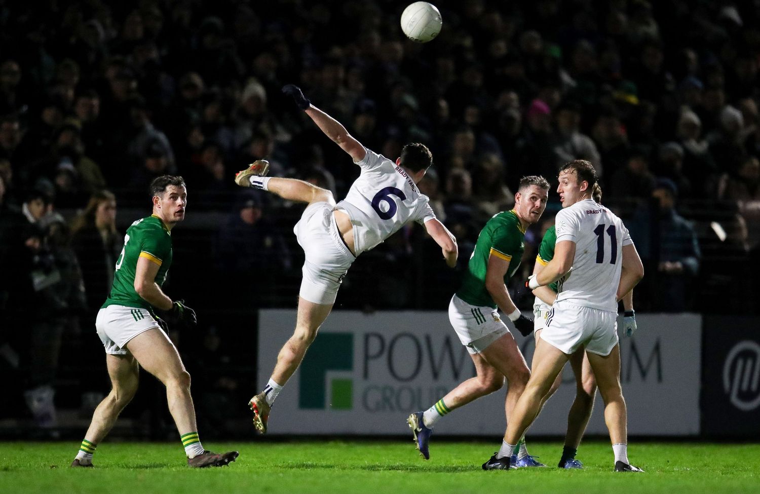 A soccer player performs an acrobatic overhead kick during a match between teams in white and green jerseys.