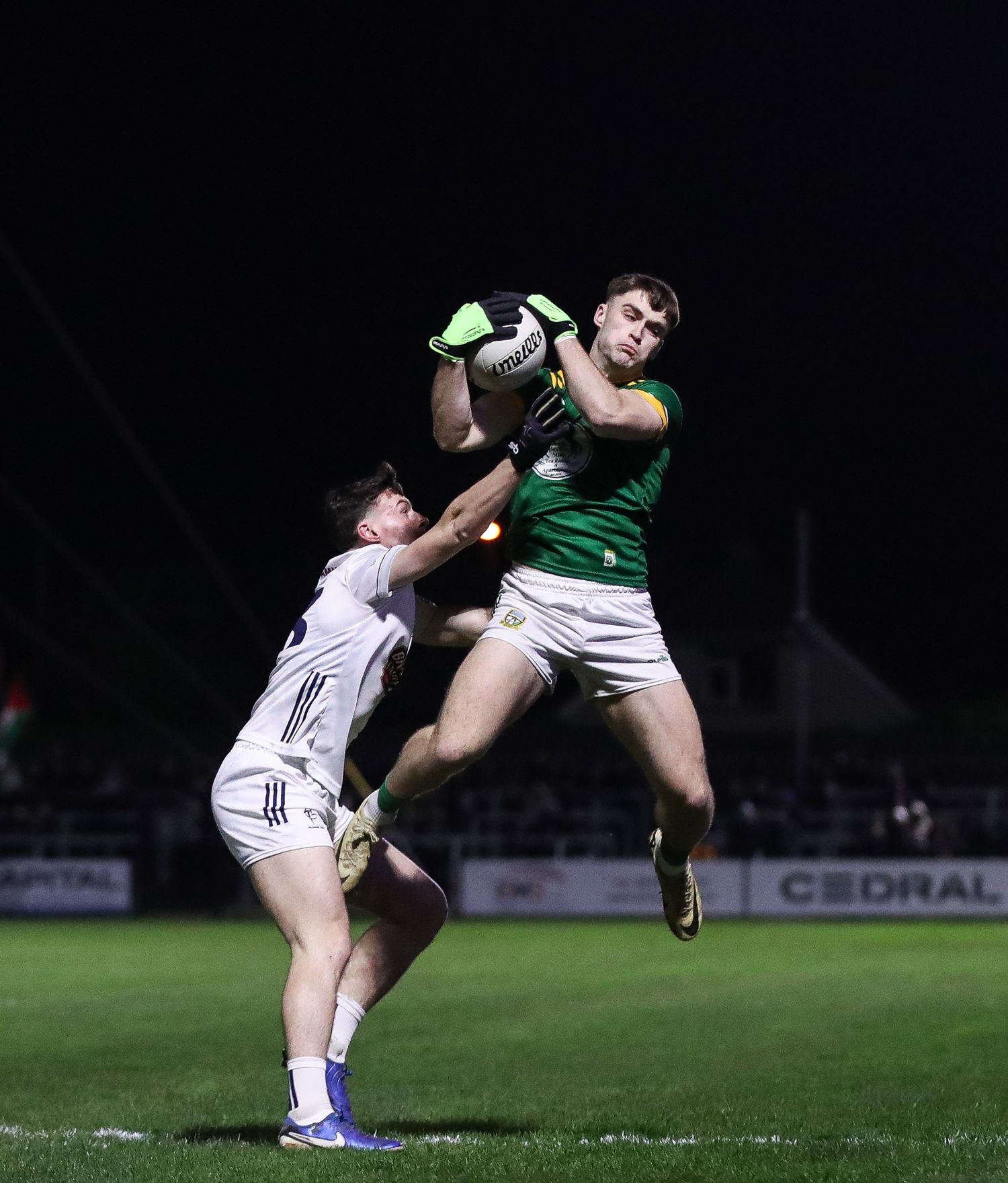 Two football players collide mid-air while competing for possession during a nighttime match.