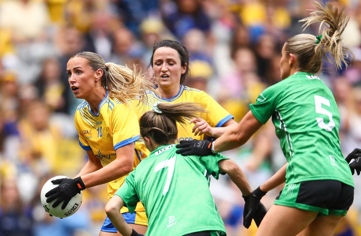 Female Gaelic football players compete in an intense match, wearing blue and yellow jerseys with a goalkeeper in green.