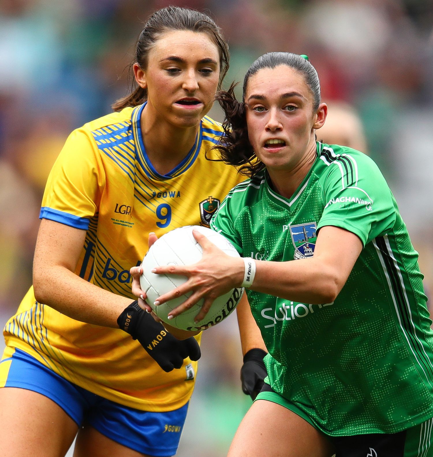Two female Gaelic football players compete for the ball during a match, wearing yellow and green jerseys.