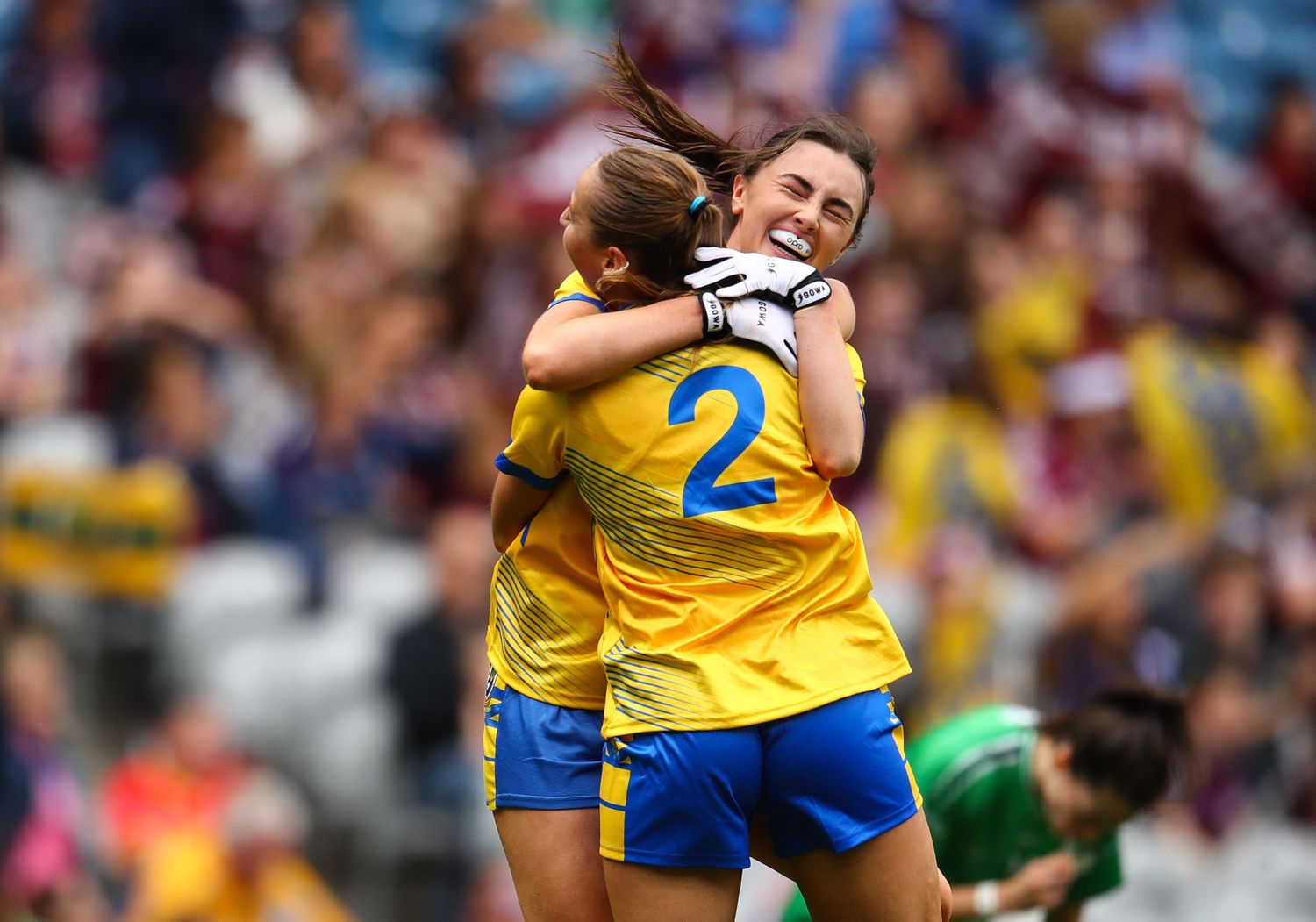Two Roscommon GAA players in yellow jerseys celebrate with a hug on the pitch during a Ladies football match.