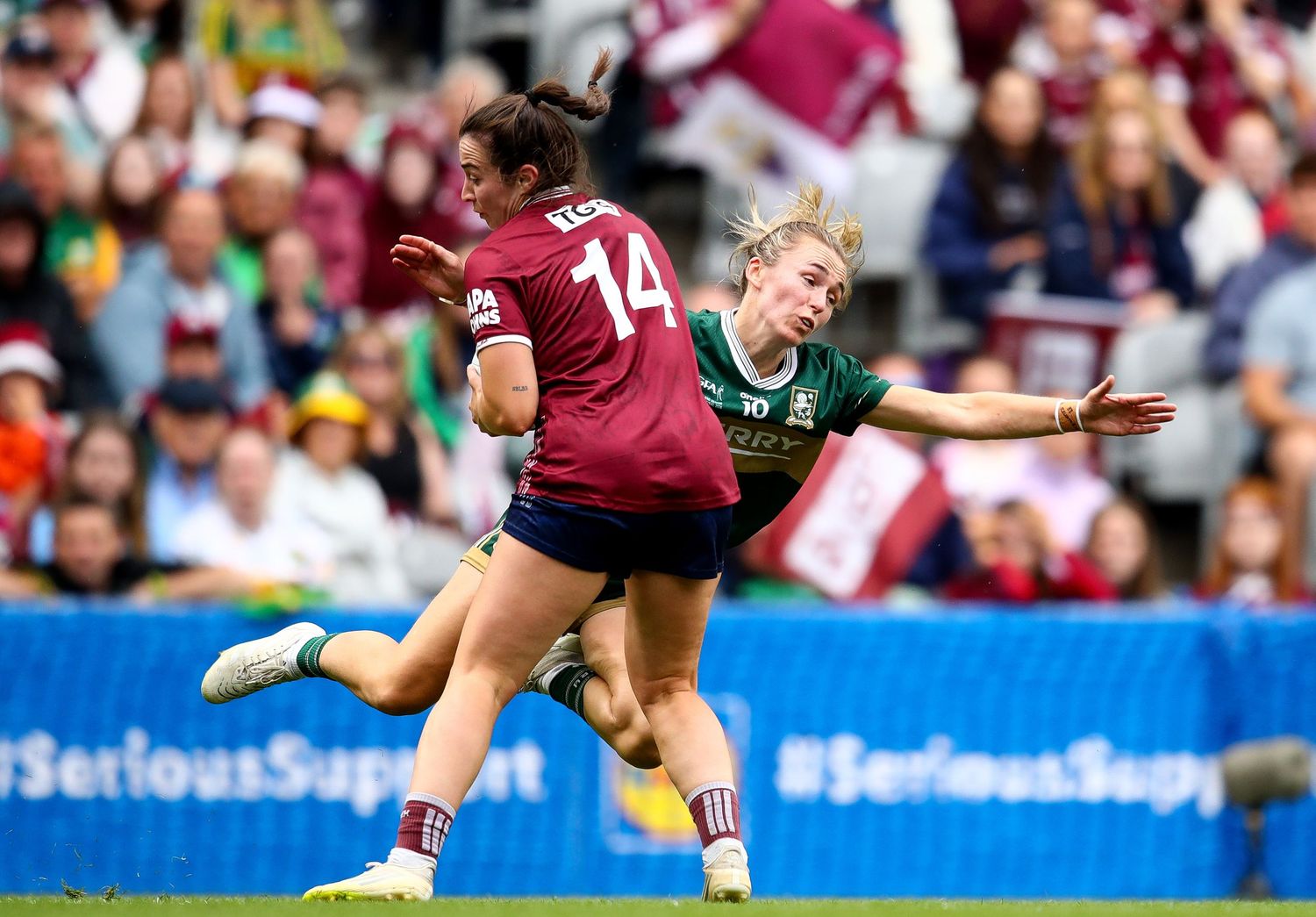 Two female Gaelic football players compete intensely during a match, wearing maroon and green jerseys on a sunny day.