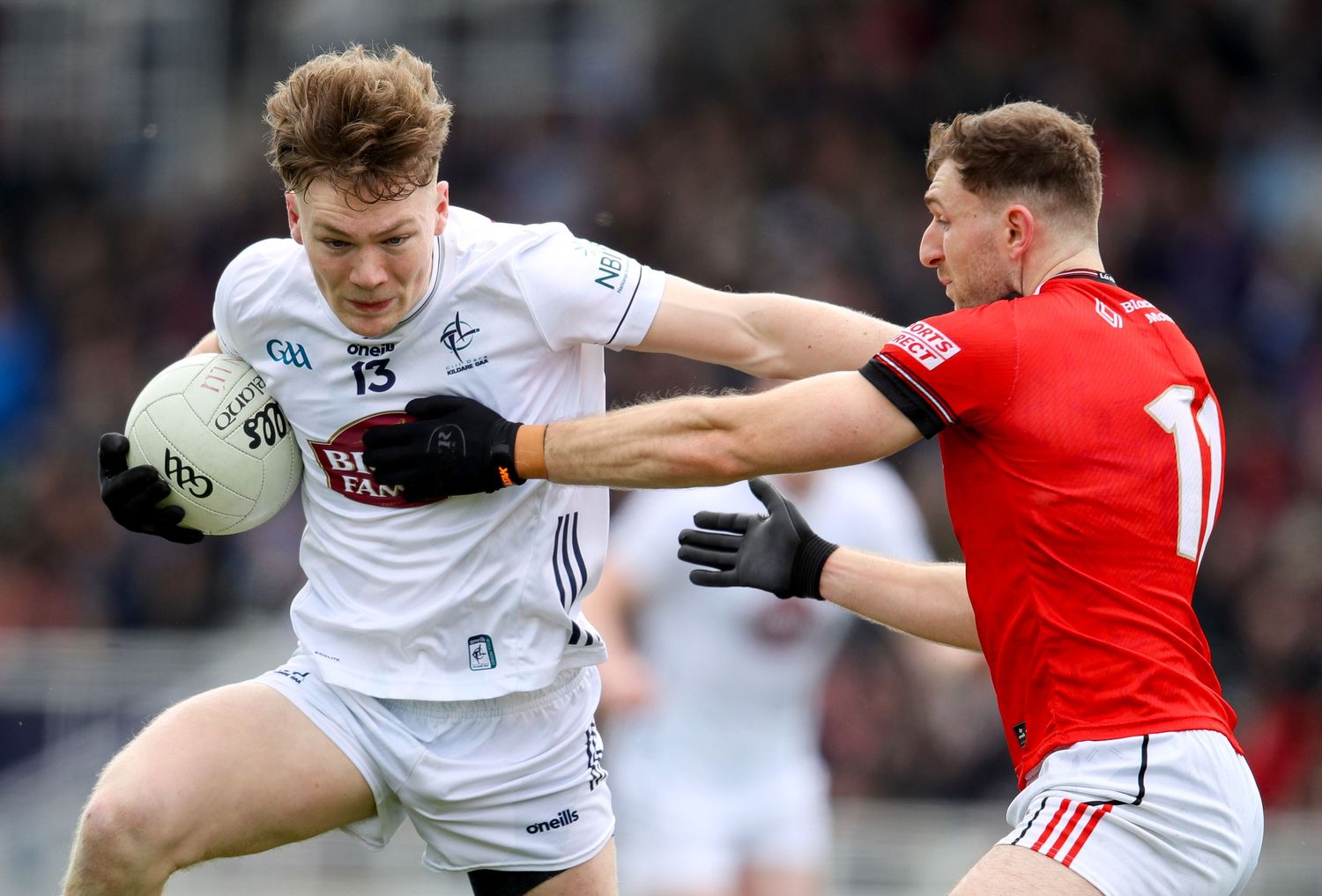 Two Gaelic football players compete for the ball during an intense match, one in white and one in red jersey.