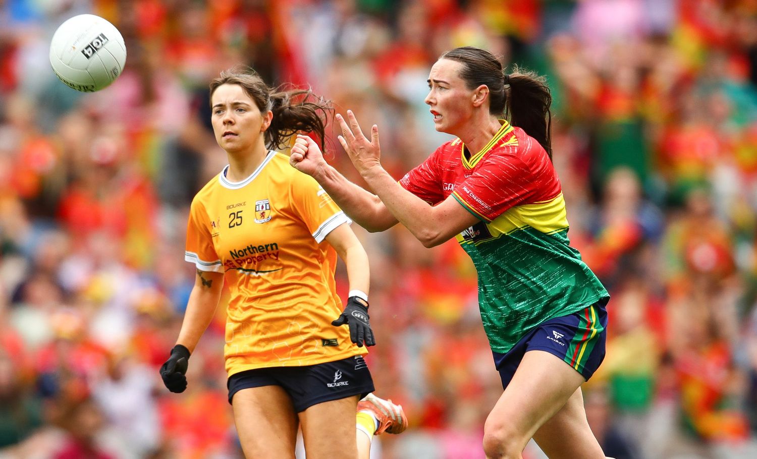 Two female Gaelic football players compete for the ball during a match, wearing Antrim and Carlow jerseys.