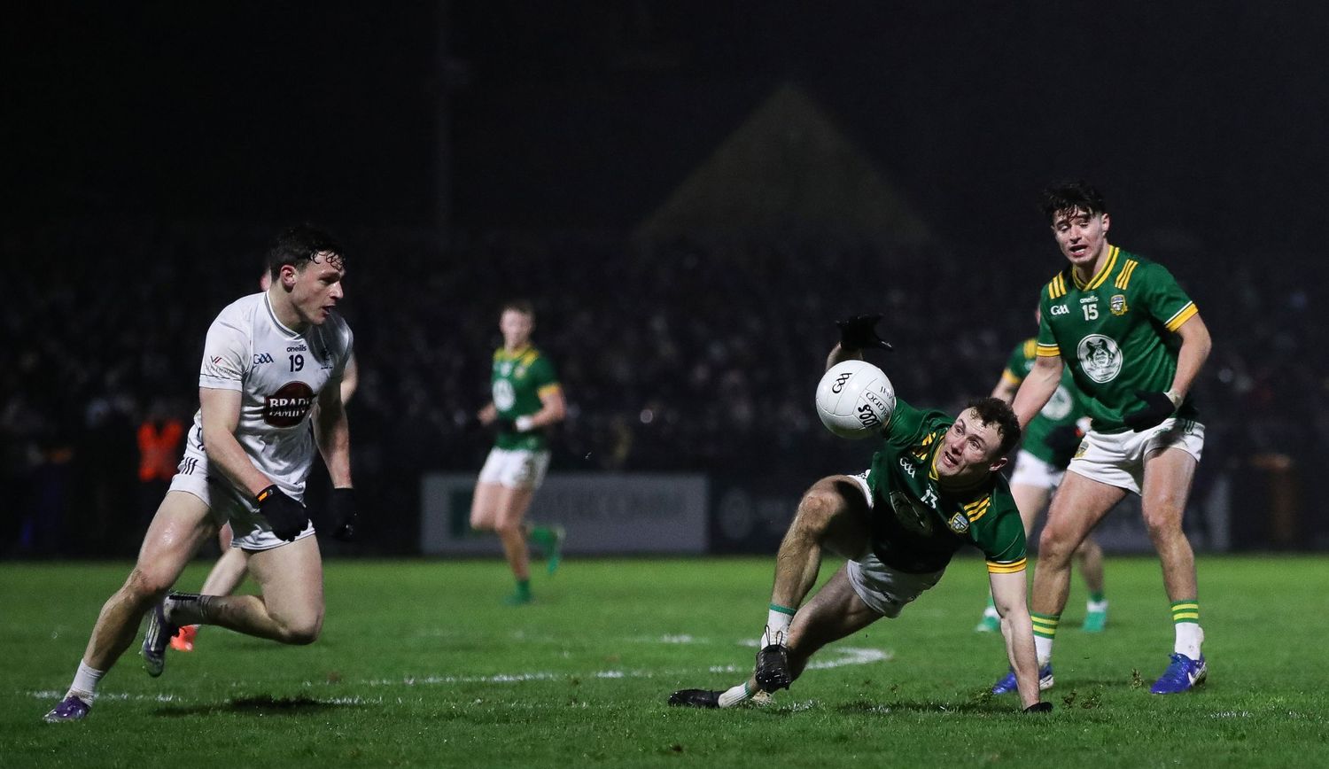 Gaelic football players from opposing teams dive for the ball during a nighttime match under floodlights.