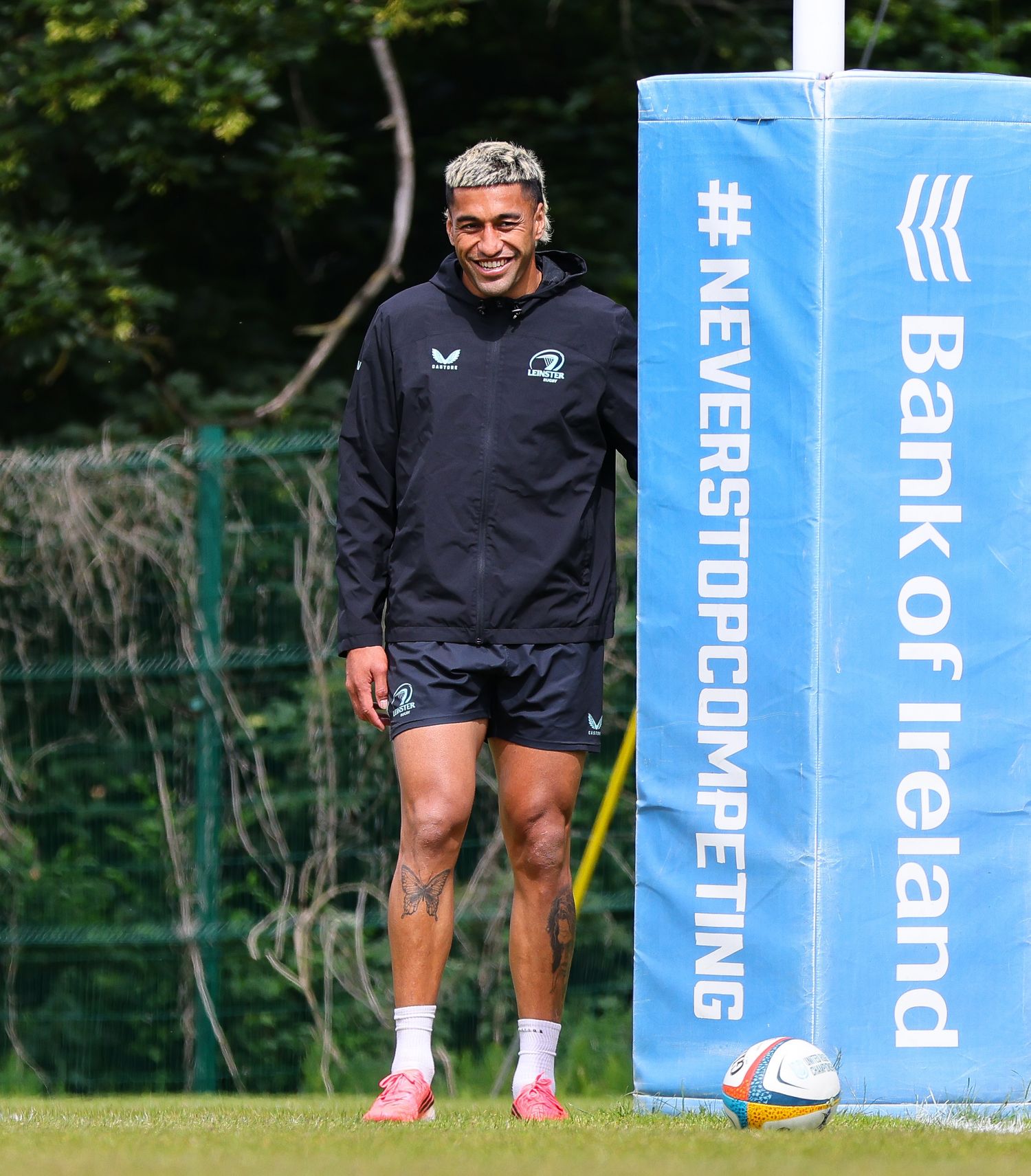 Rugby player smiling at Leinster training session beside Bank of Ireland #NeverStopCompeting banner on sunny day.
