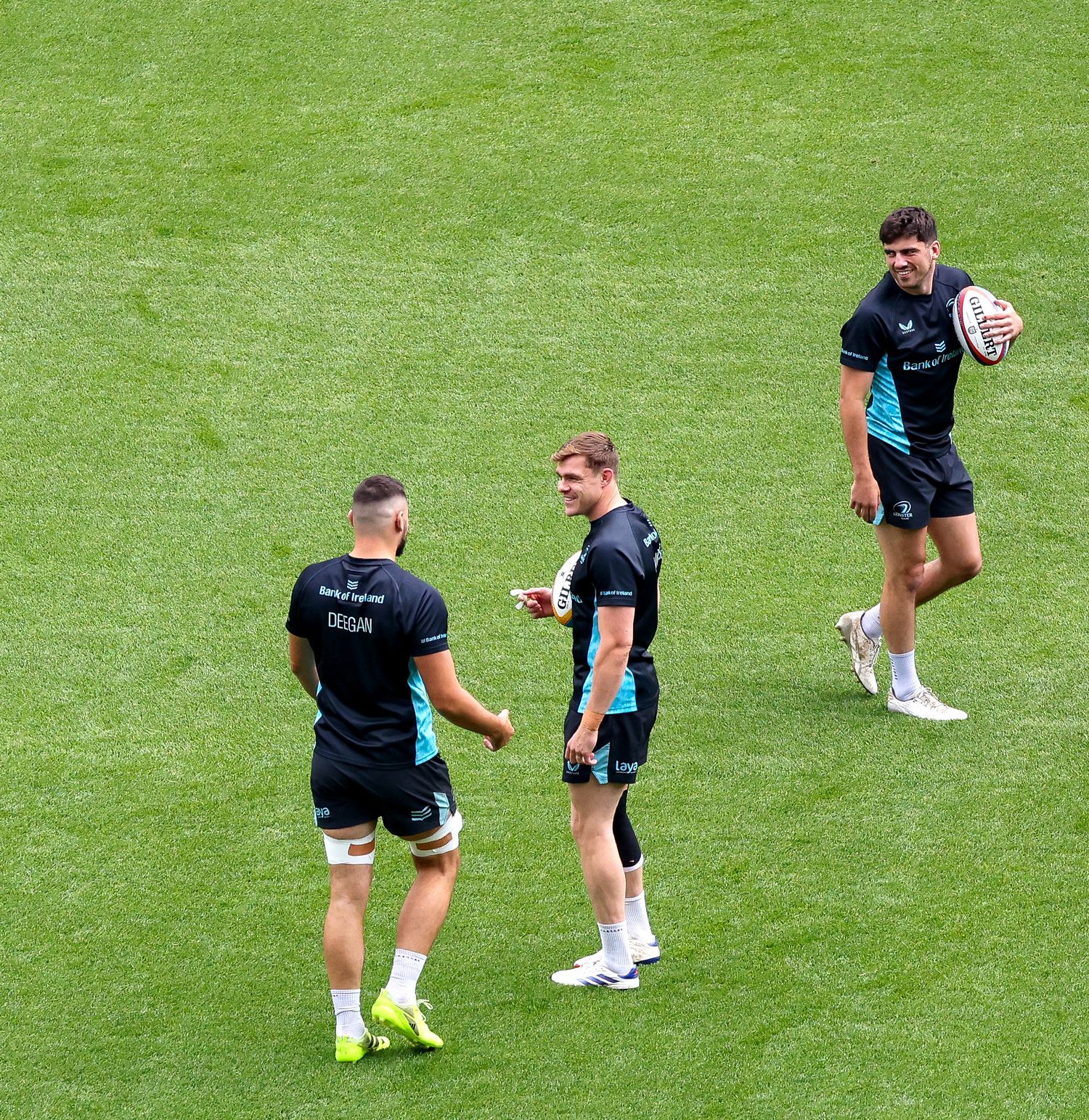Three rugby players in dark uniforms practice on a green field, with one player holding a rugby ball.