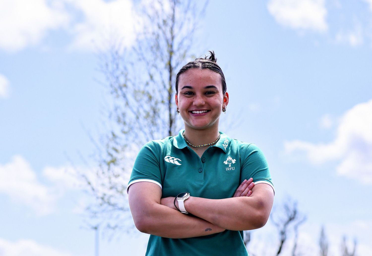 Woman in green Ireland rugby polo shirt smiling with arms crossed outdoors on a sunny day.