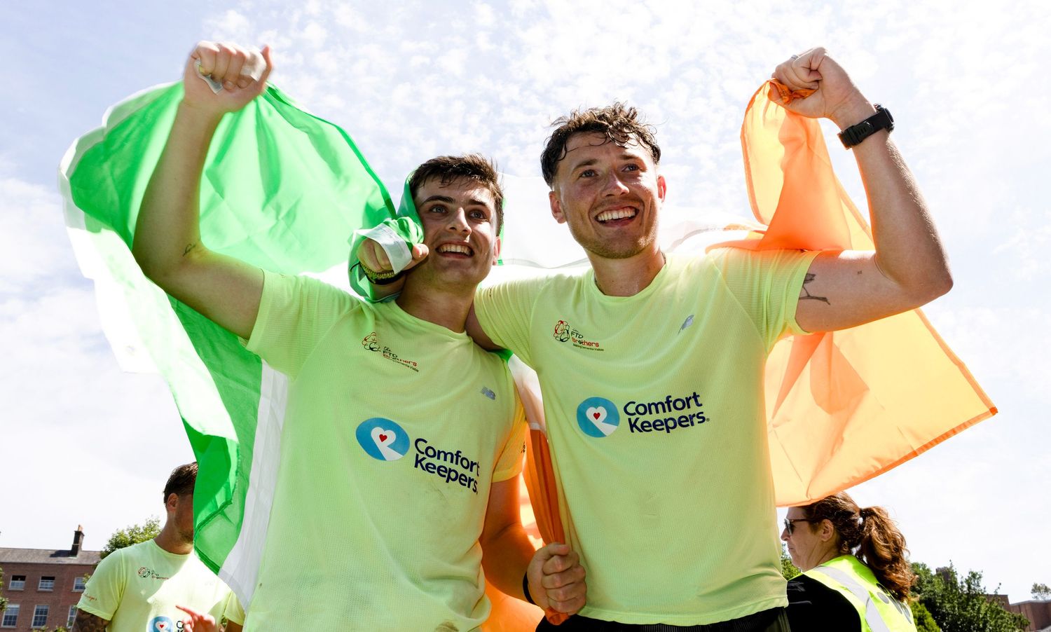 Two men in Comfort Keepers shirts celebrate outdoors, holding a green and orange flag with arms raised in victory.