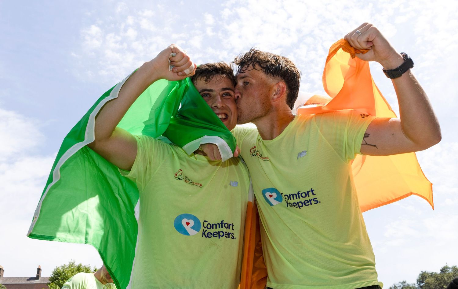 Two young men in yellow Comfort Keepers shirts celebrate outdoors, holding an Irish tricolor flag with raised fists.