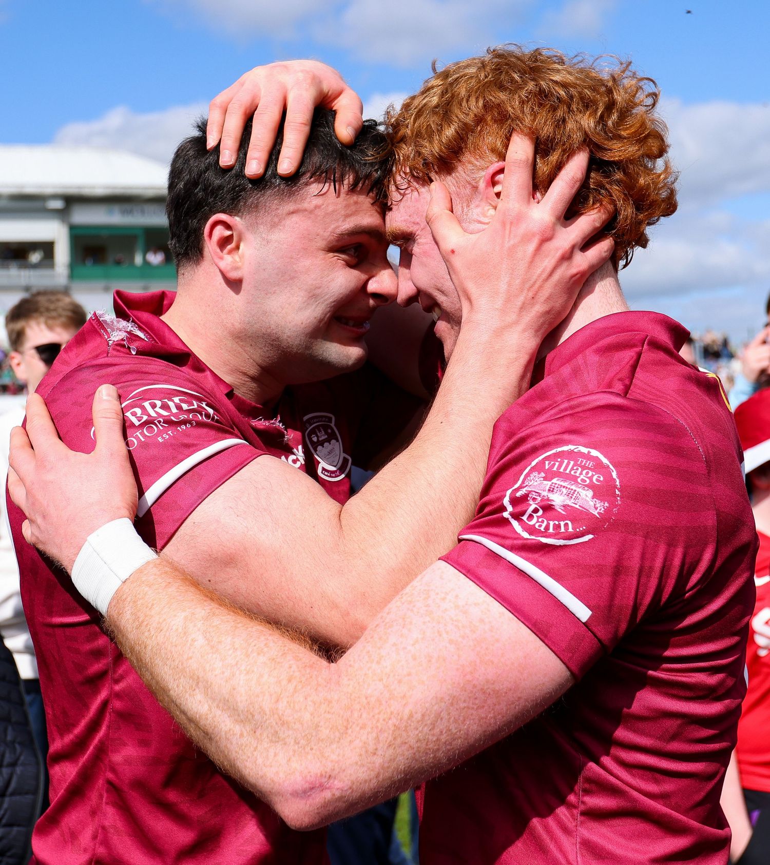 Two rugby players in maroon jerseys share an emotional embrace on the field after a match.