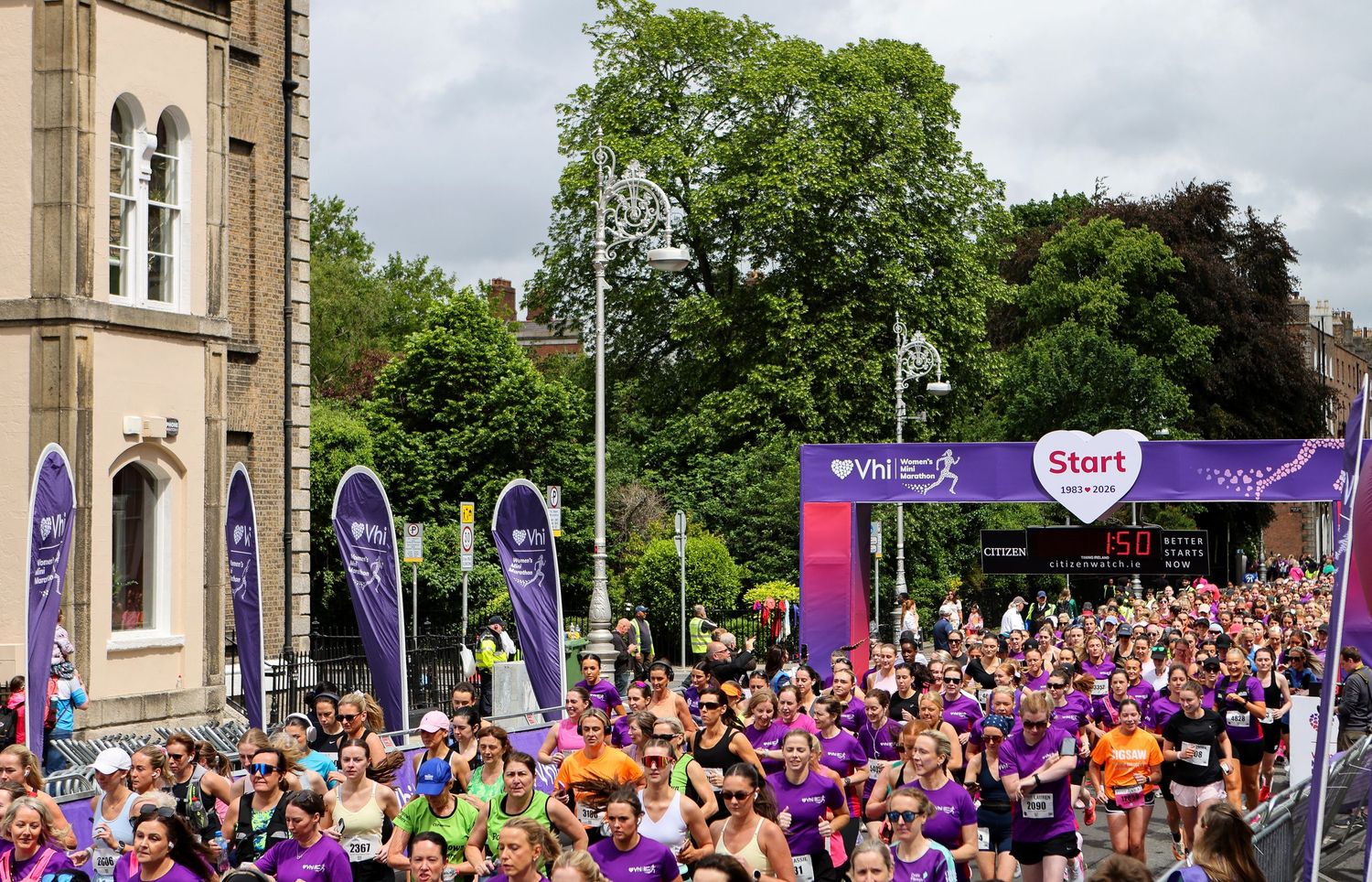 Crowds of runners in purple shirts gather at the start line of an outdoor charity fun run event on a sunny day.