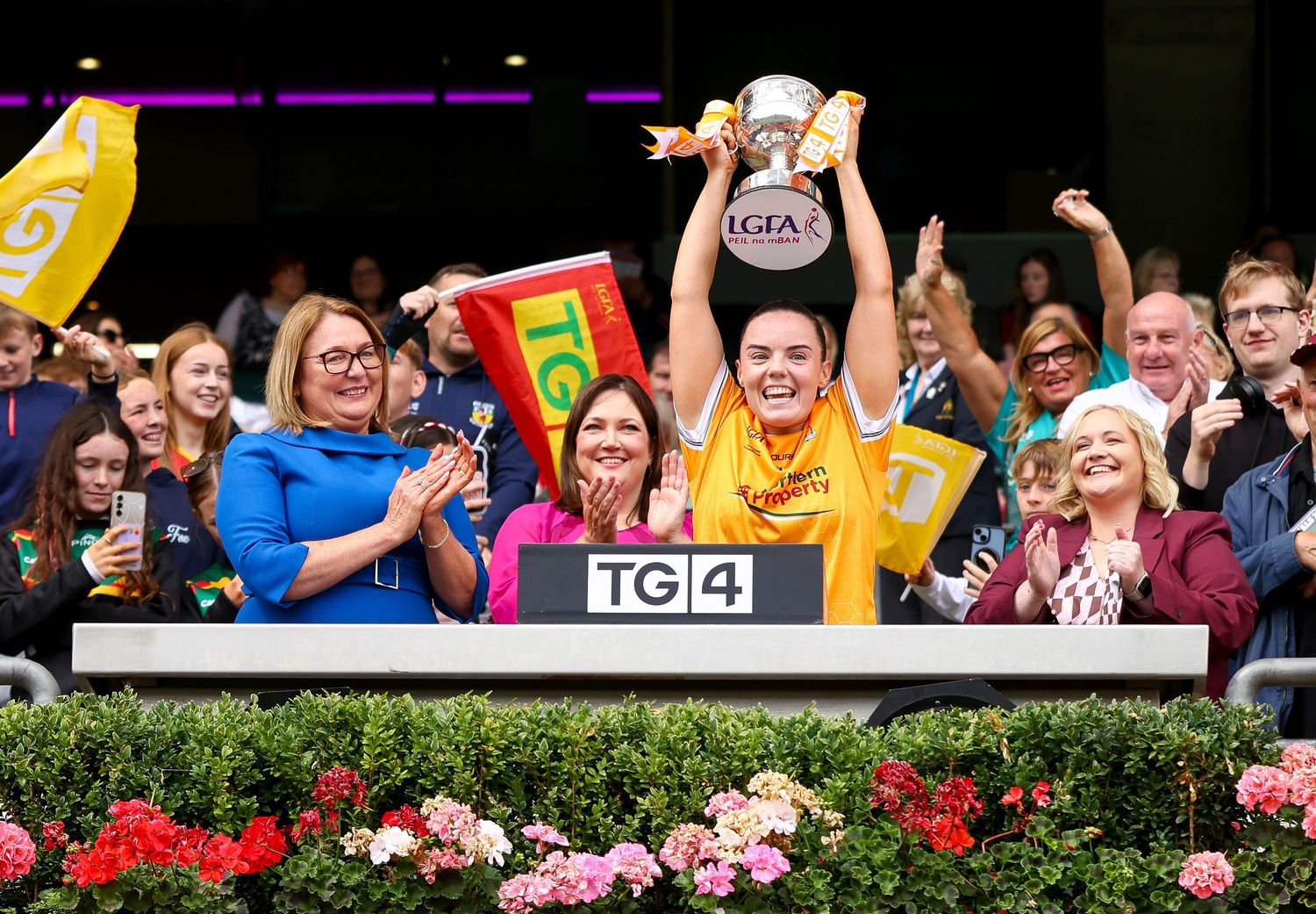 A woman triumphantly lifts a trophy at a LGFA event, surrounded by cheering fans and colorful flags in the stands.