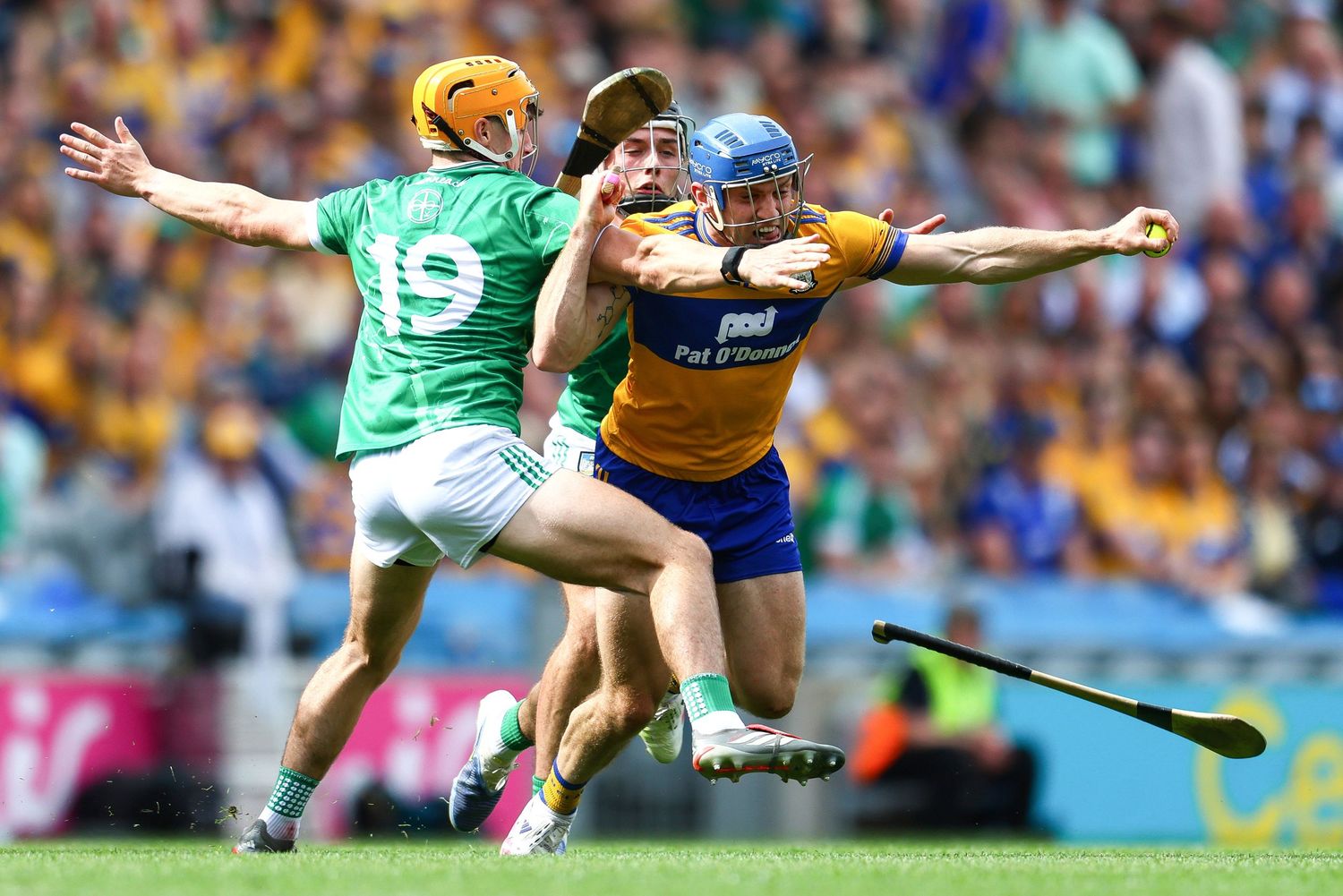Two hurling players compete fiercely during a GAA match, with a hurley stick flying through the air.