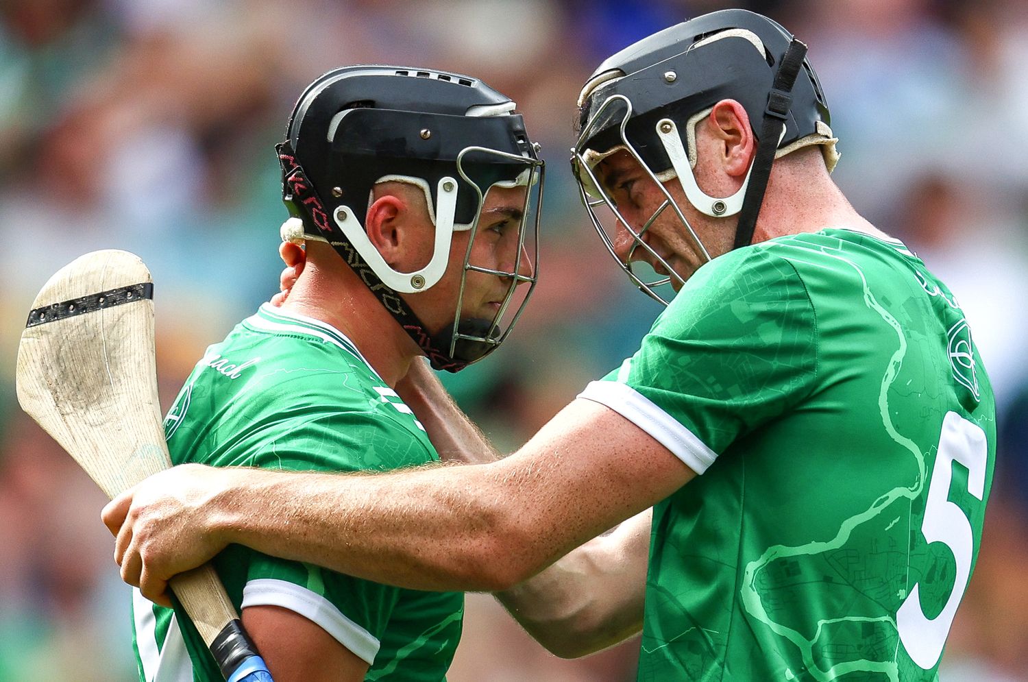 Two hurling players in green jerseys and helmets embrace during a match, one wearing number 5.