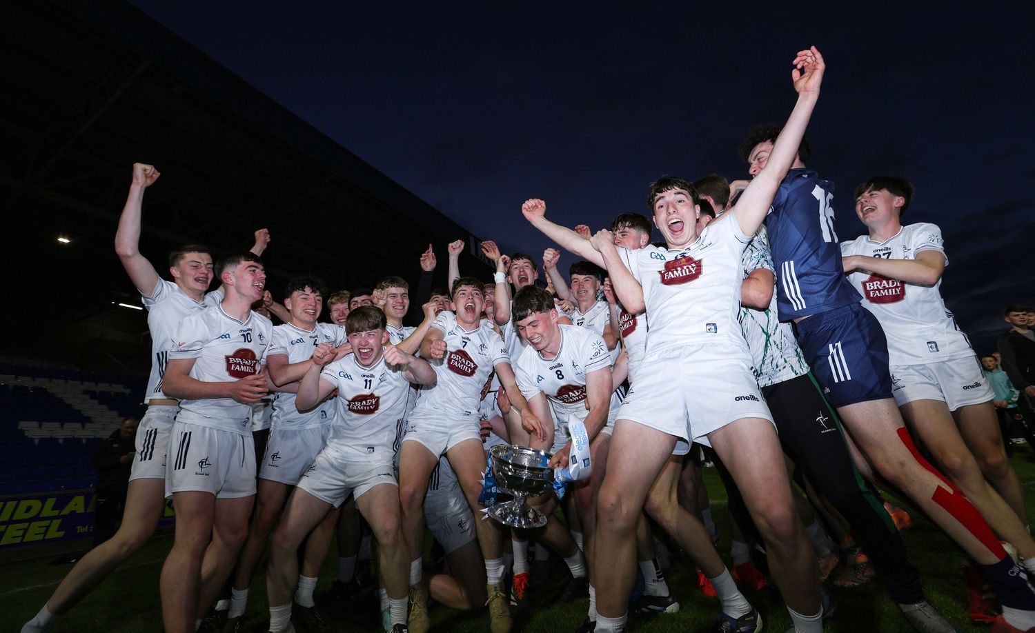 Kildare GAA team celebrating a championship victory, players cheering with trophy under floodlights.